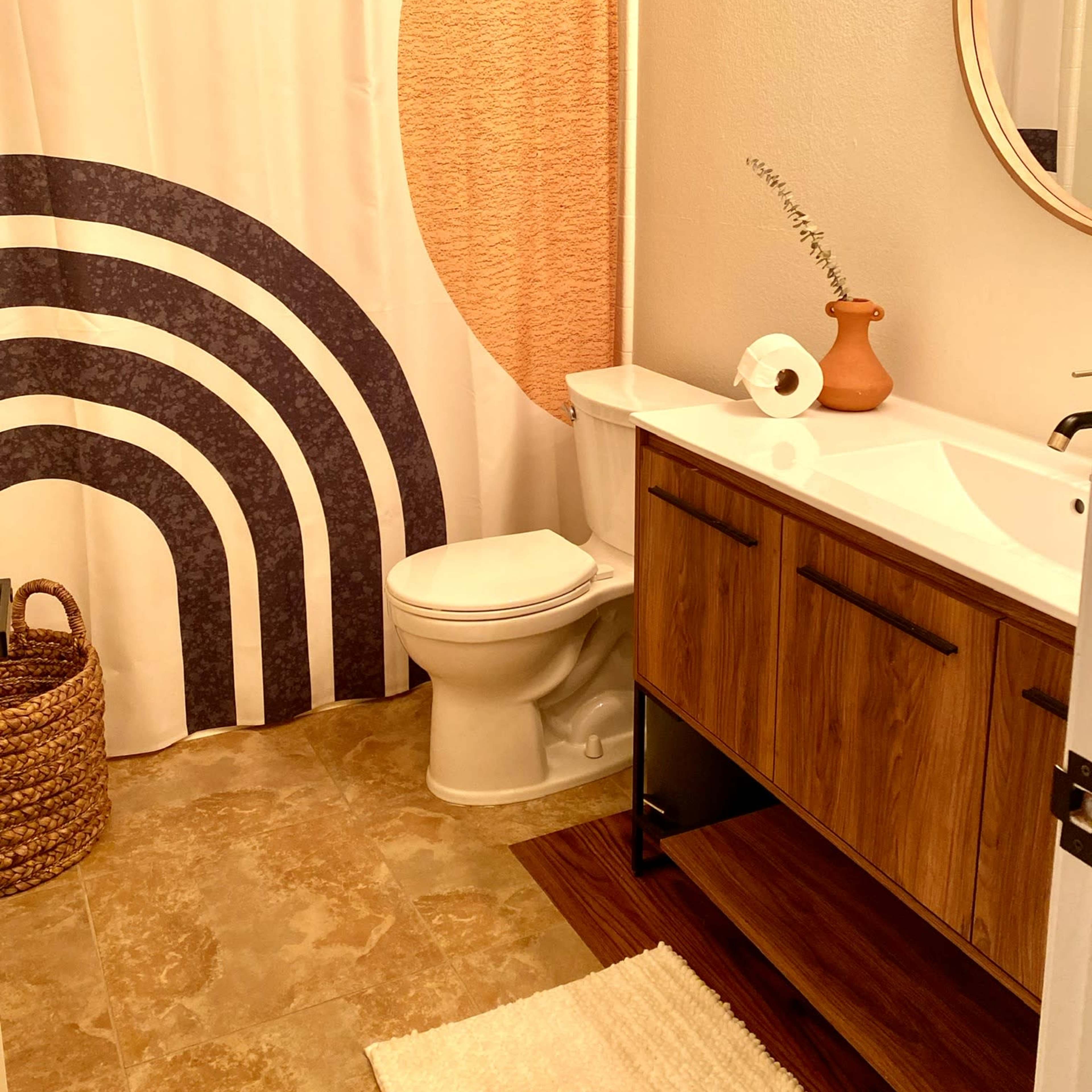A bathroom featuring a shower curtain with a geometric design, a wooden vanity with a circular mirror, a toilet, and a woven basket in the corner.