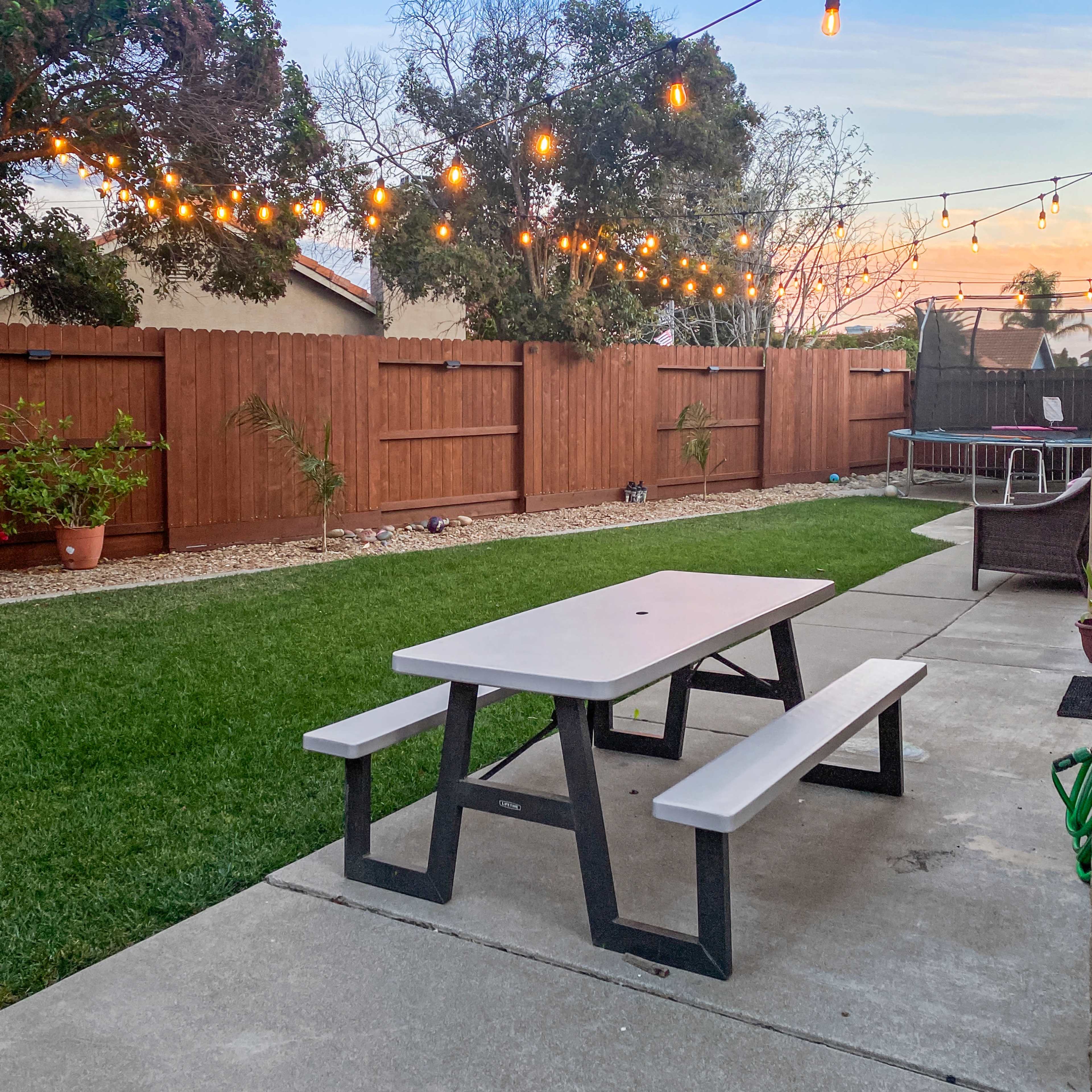 A backyard with a concrete patio, a picnic table, and warm string lights hanging above a grassy area and wooden fence.