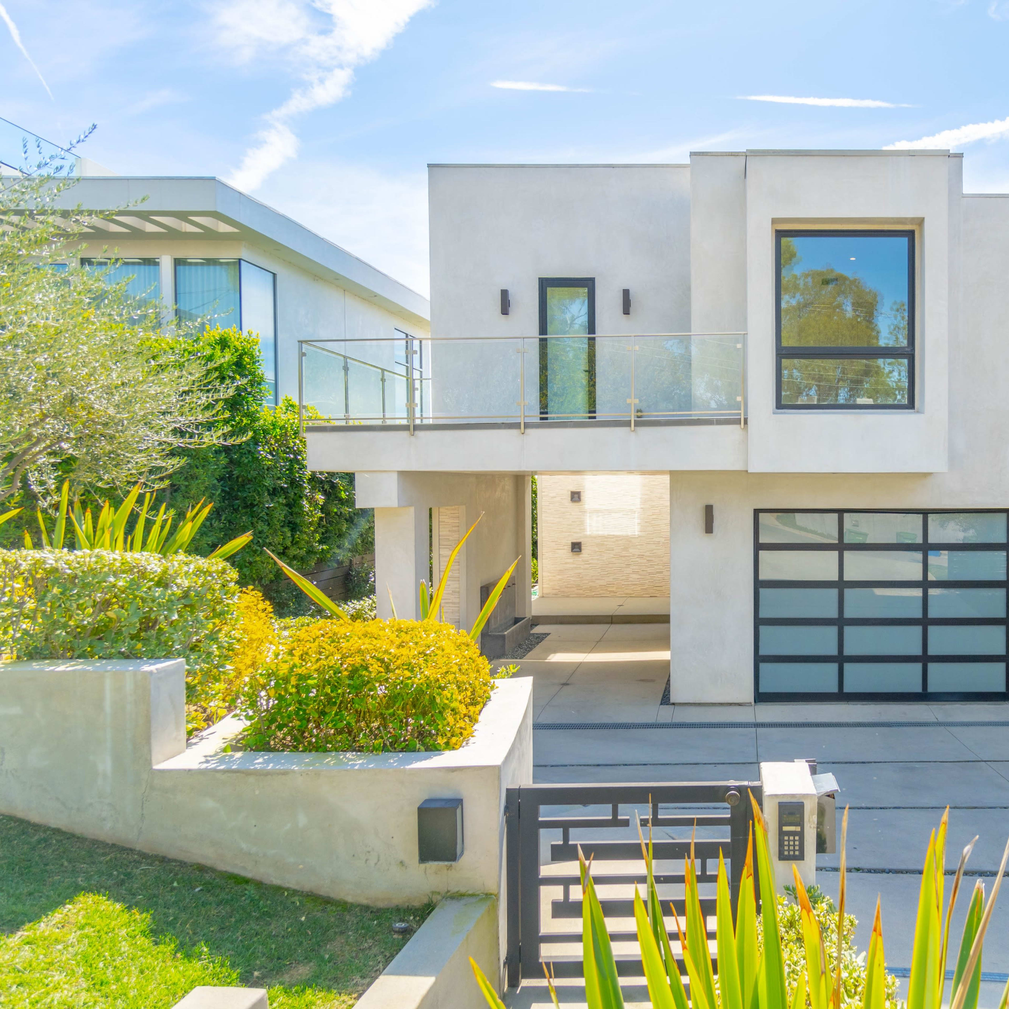 A modern two-story house with a glass-railed balcony, a large garage door, and landscaped greenery in the front yard.