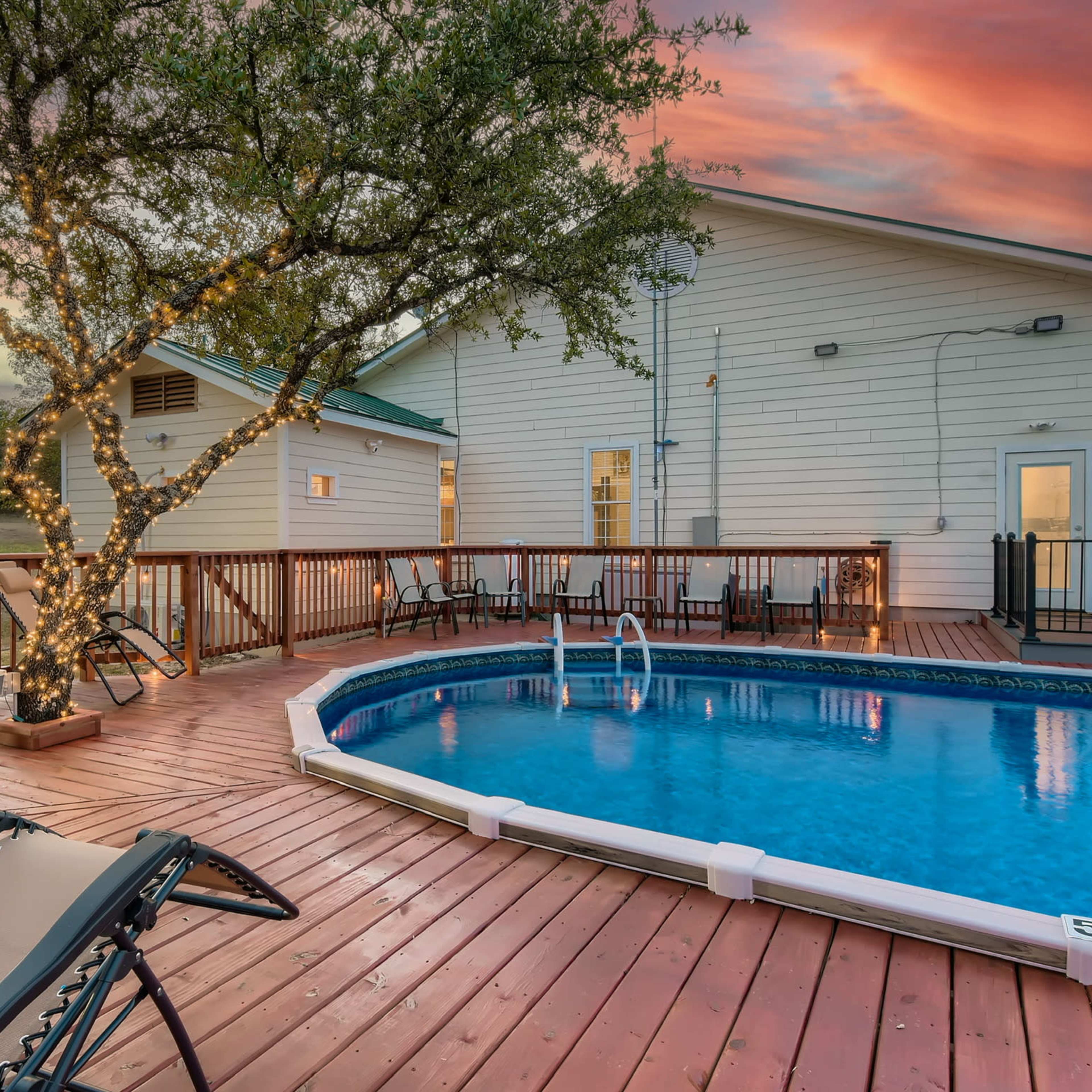 A wooden deck with lounge chairs surrounding a rectangular swimming pool, illuminated by string lights on a nearby tree, under a colorful sunset sky.