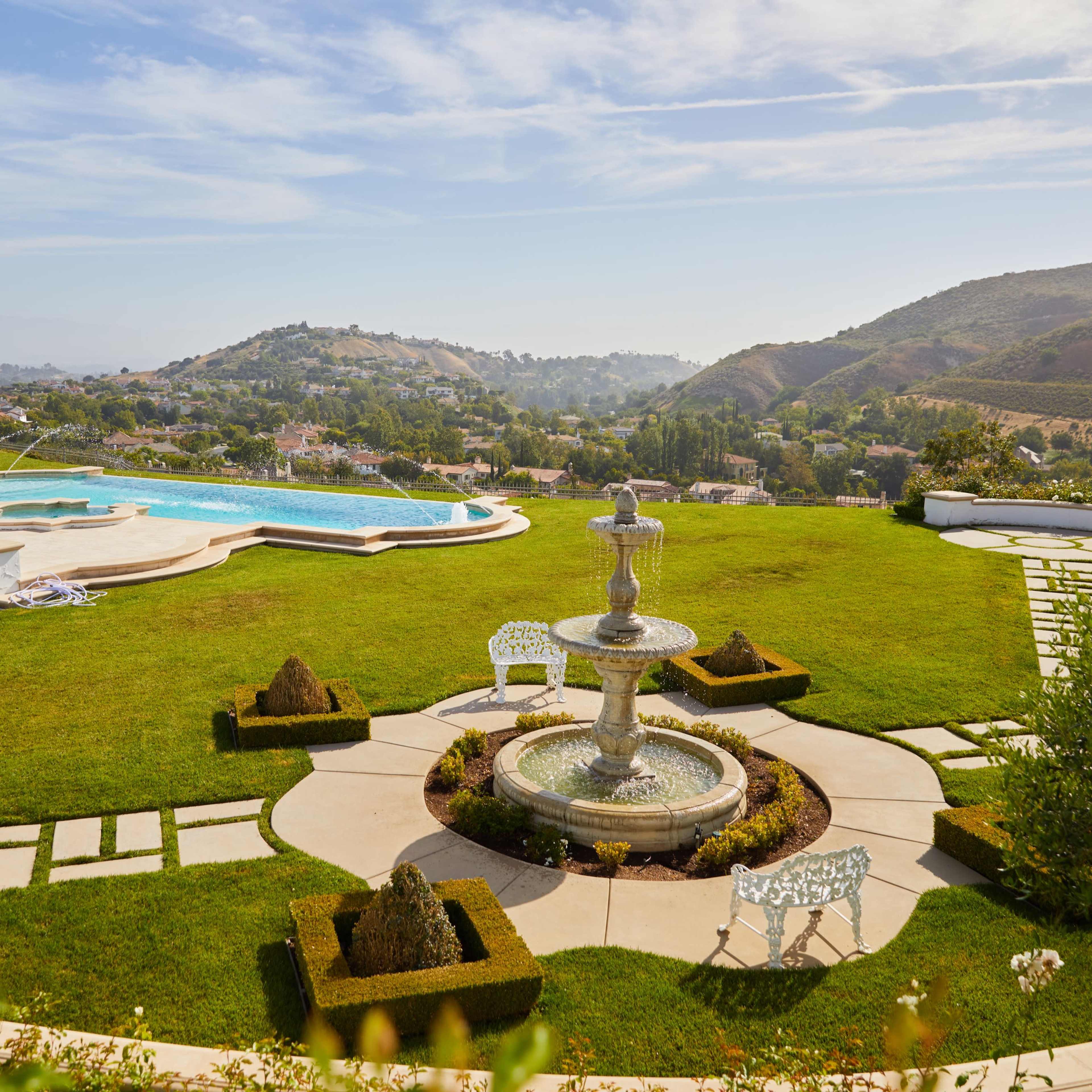 A sprawling garden with a swimming pool, a fountain, and landscaped pathways, set against rolling hills in the background.