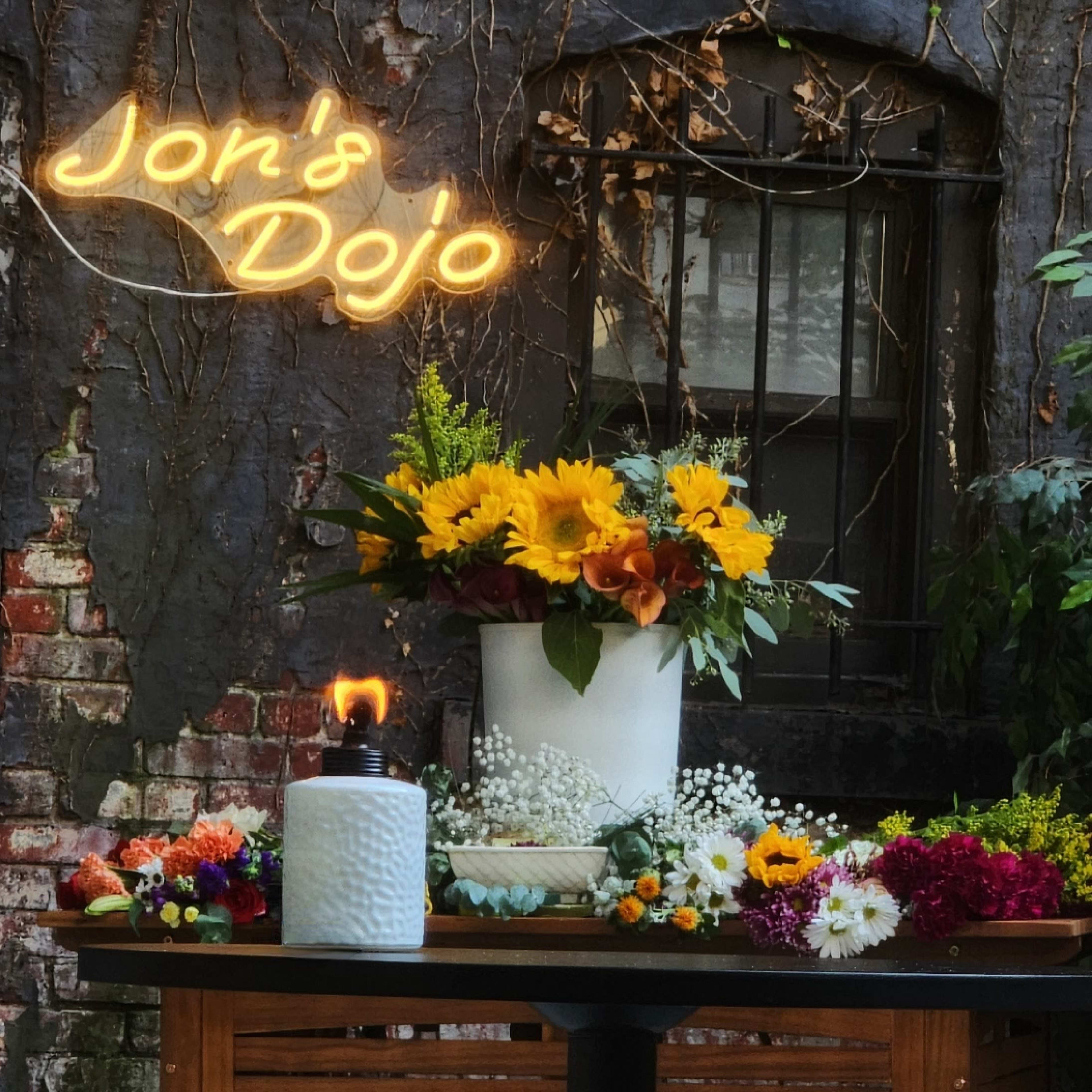 A wooden table adorned with various flowers and a candle is set against a brick wall with a neon sign reading "Jon's Dojo."