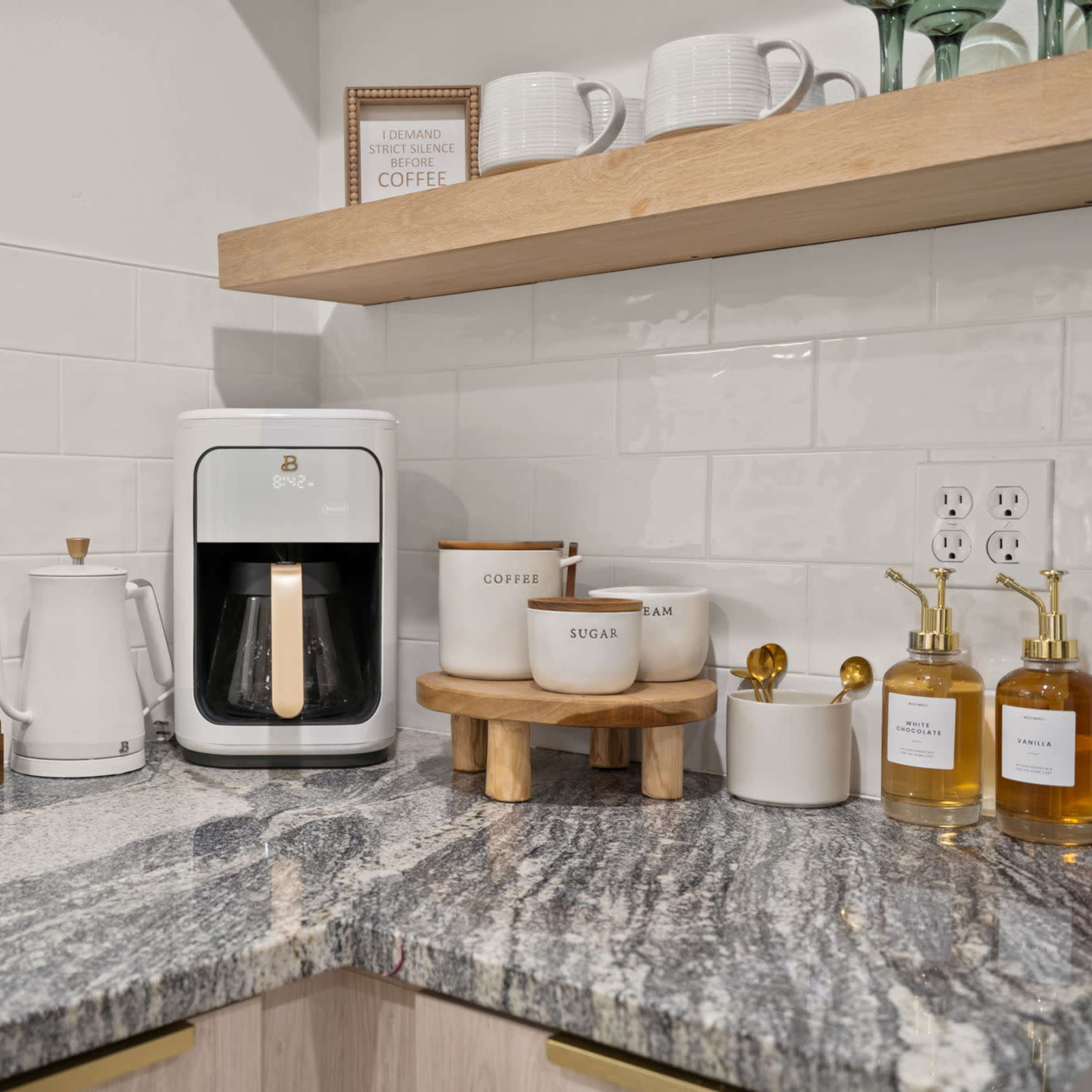 A kitchen countertop featuring a coffee maker, coffee pot, jars labeled for coffee and sugar, and a set of mugs on a wooden shelf.
