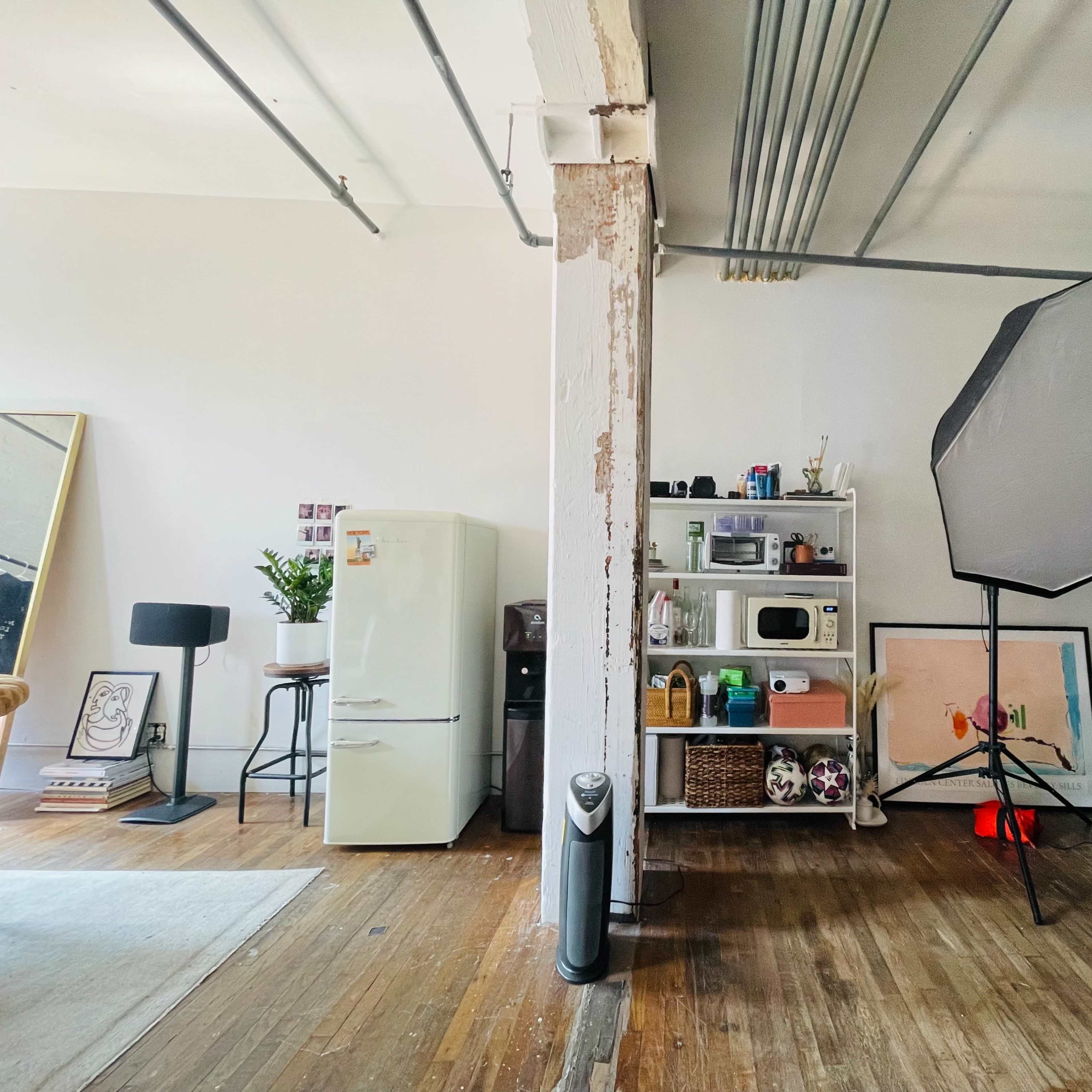 An interior space with a green couch, a shelf filled with appliances and decor, a refrigerator, and a large softbox lighting setup.