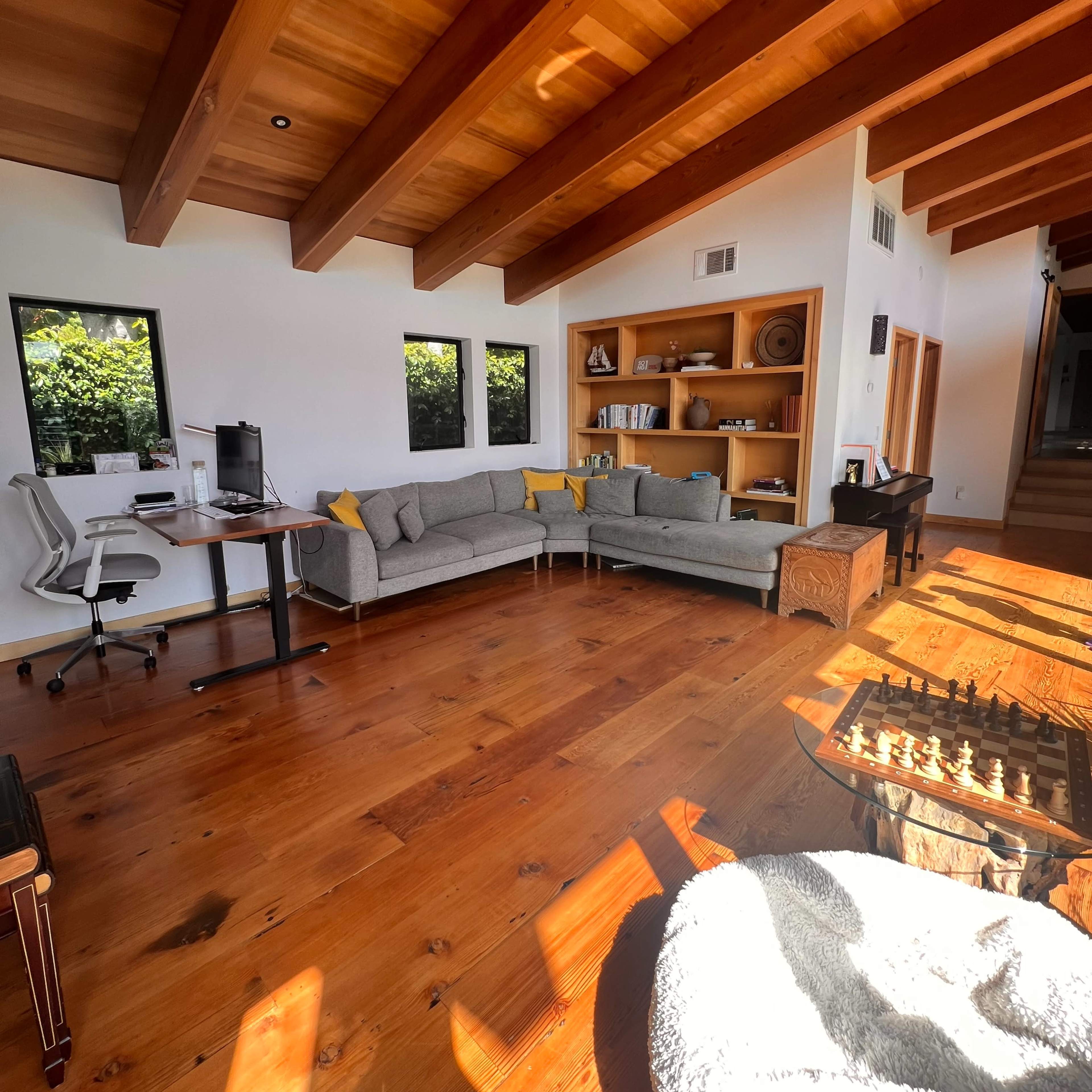 A spacious living room with wooden beams, a gray sectional sofa, a desk with a computer, and a chess table on the floor.