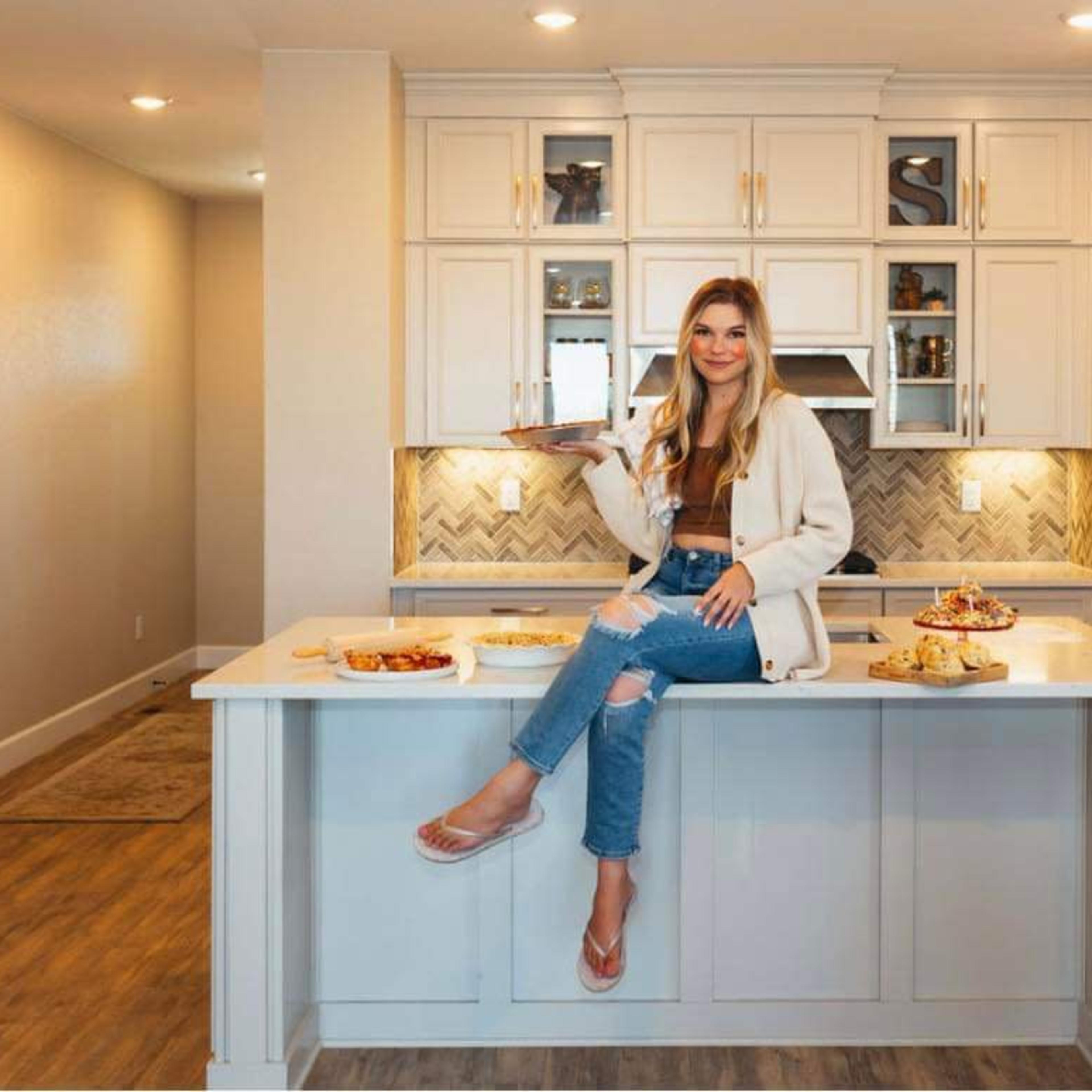 A woman sits on a kitchen island surrounded by various baked goods in a modern kitchen.