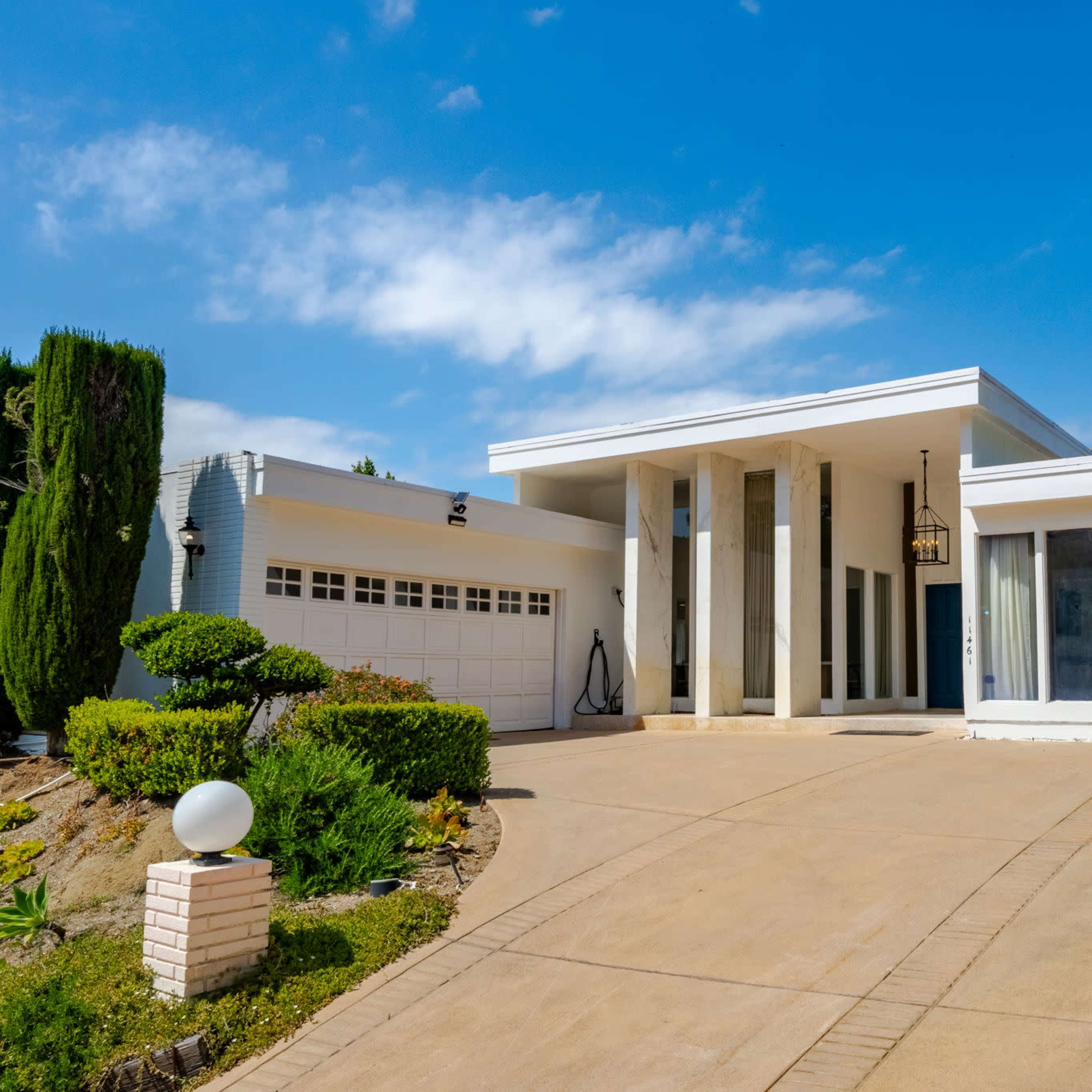 A modern, single-story house with a spacious driveway, flanked by neatly trimmed bushes and a clear blue sky.