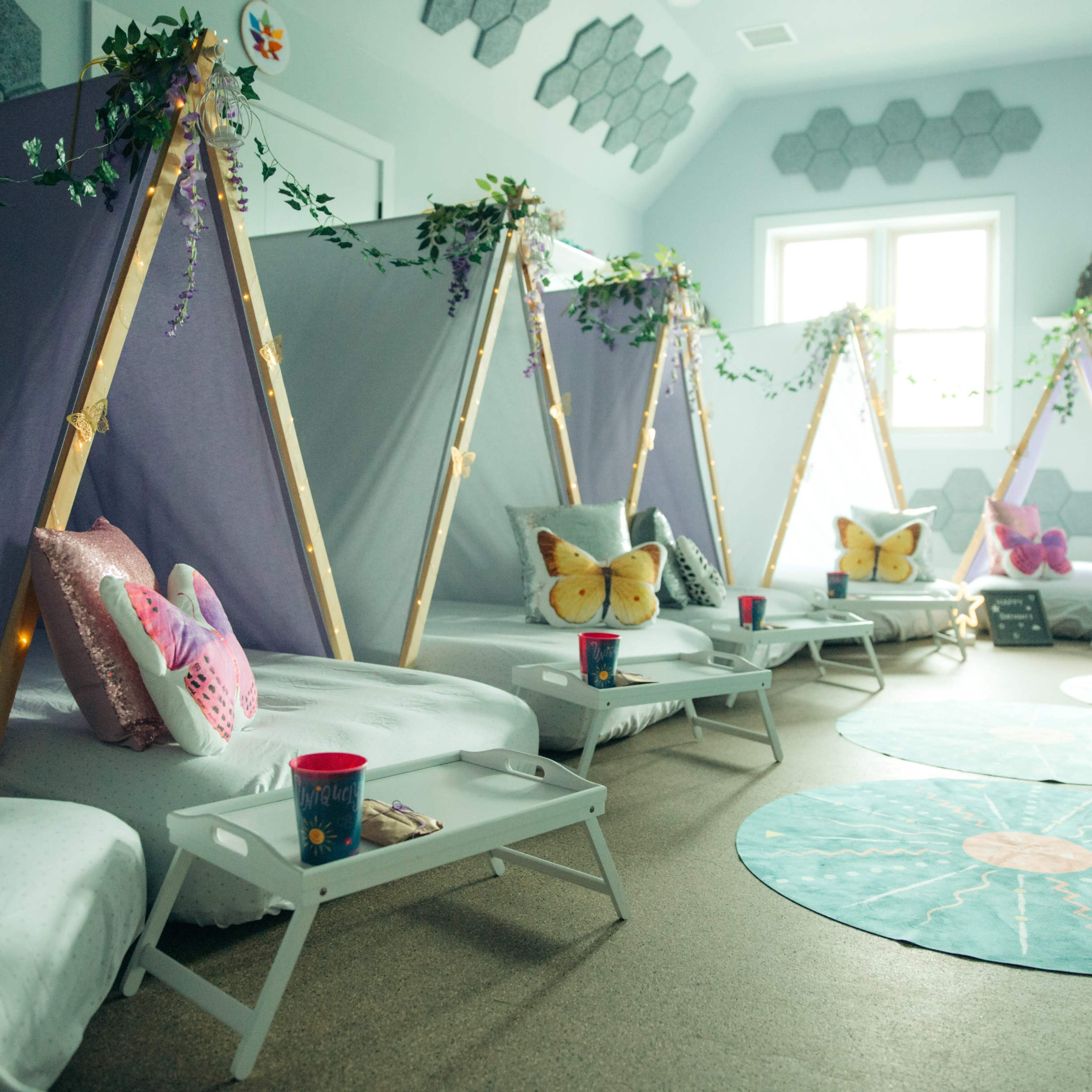 A cozy indoor setup featuring teepee-style tents, with white bedding and decorative butterfly pillows, arranged in a brightly lit room.