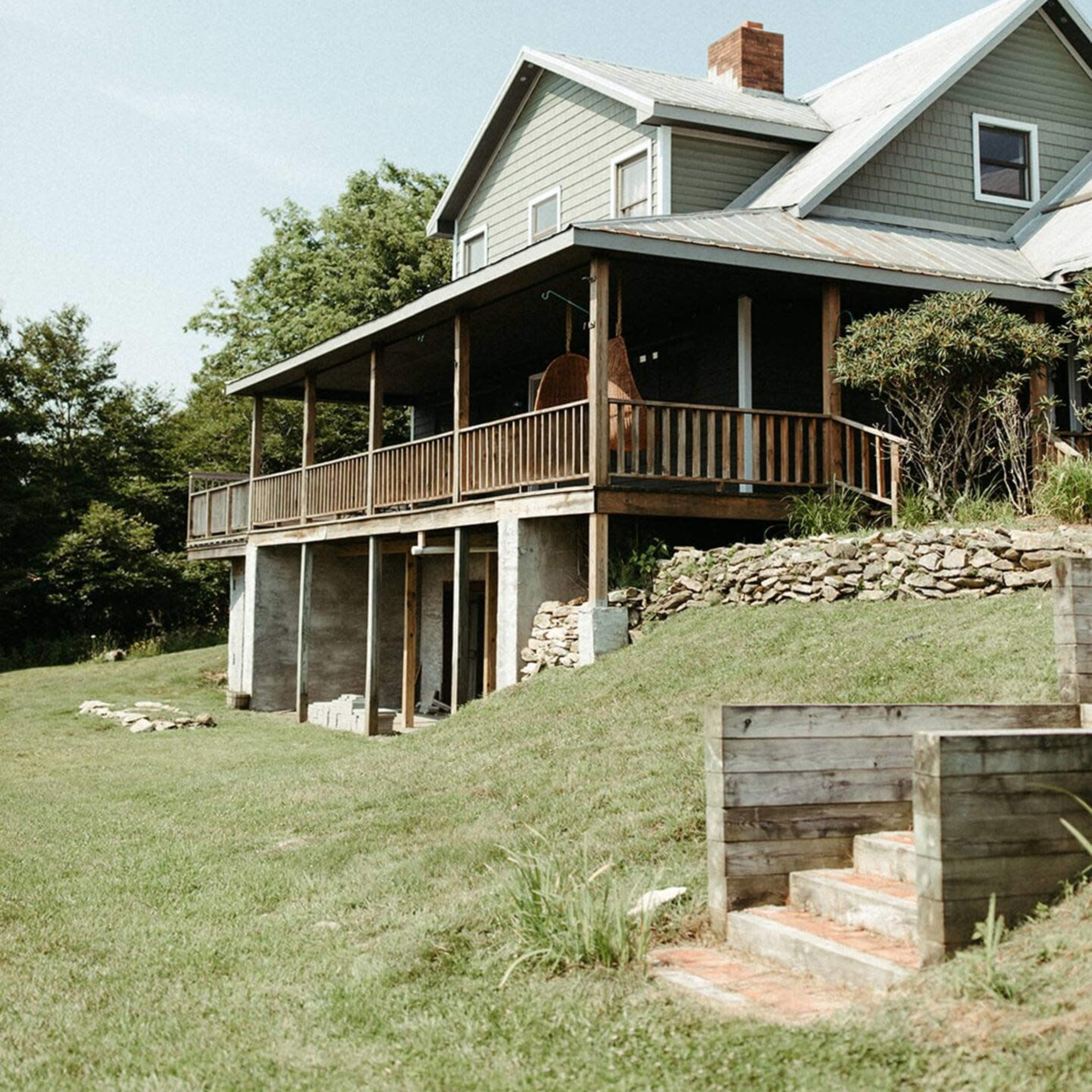 A two-story house with a wooden porch and stone foundation stands on a grassy hillside.