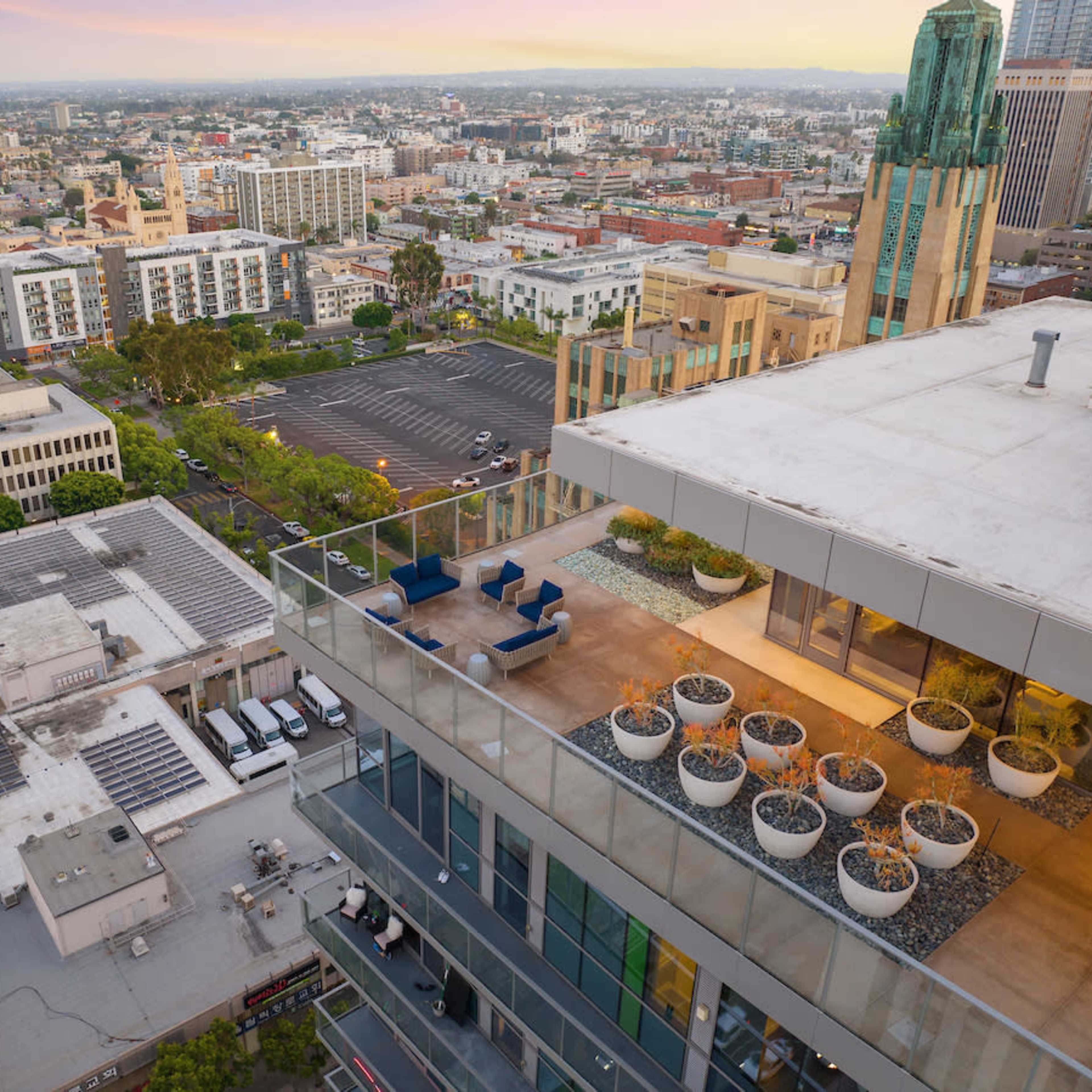 A high-rise rooftop terrace with modern furniture and planters, overlooking a cityscape with various buildings and a clear horizon.