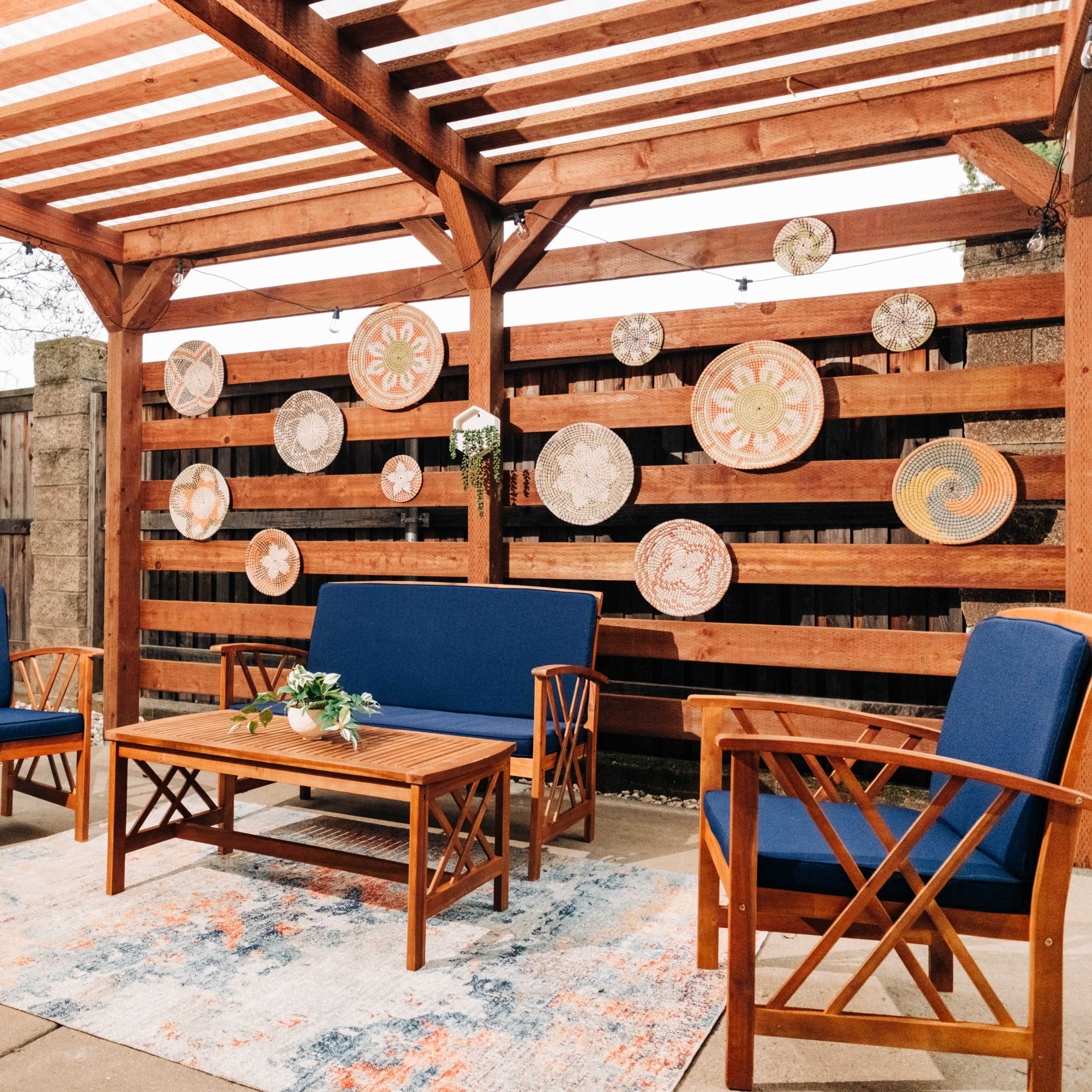 A wooden pergola with a slatted roof shelters a seating area featuring two blue chairs and a table, surrounded by decorative plates mounted on a wooden fence.