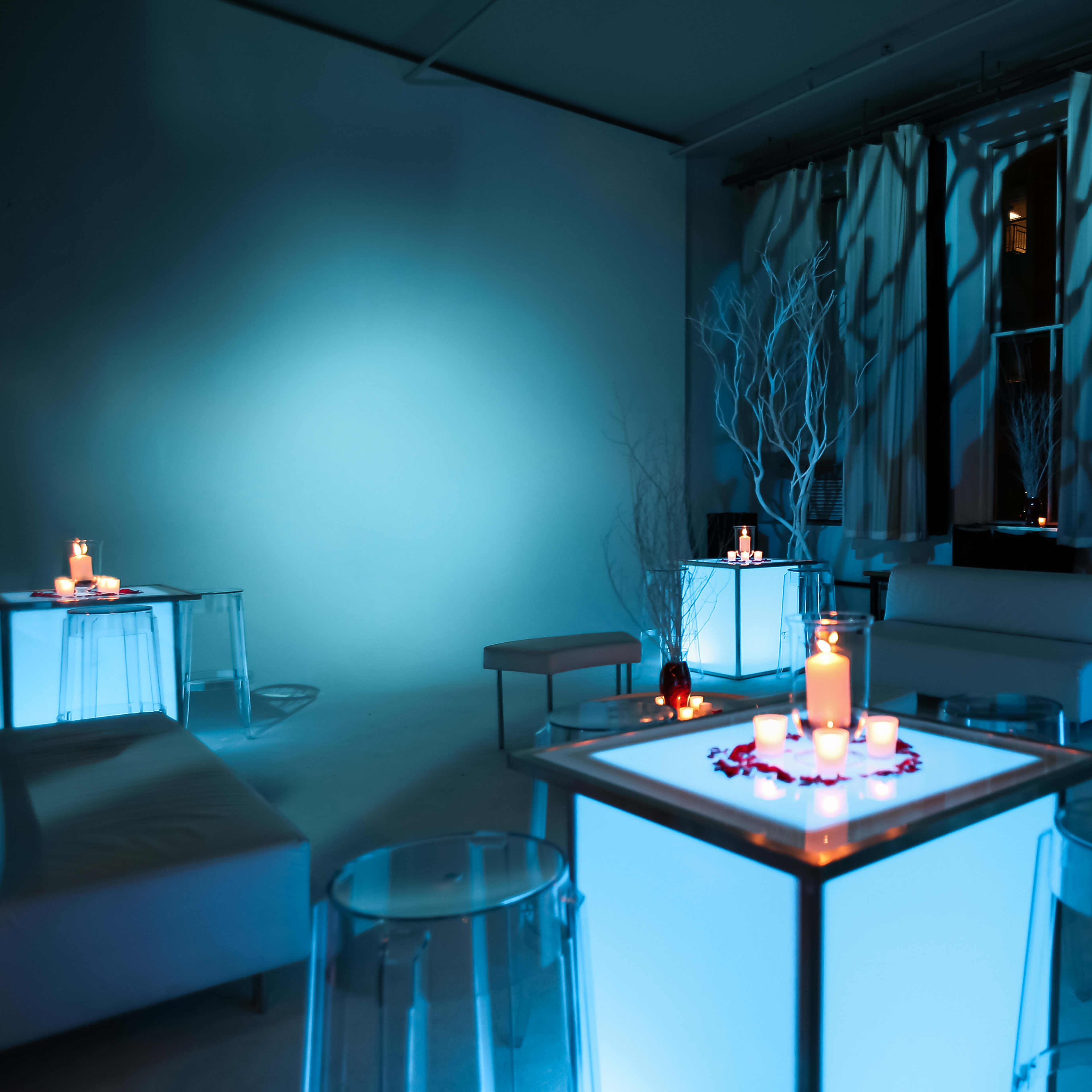 The scene features a dimly lit room with glowing blue tables adorned with candles and decorative elements, complemented by light filtering through sheer curtains.