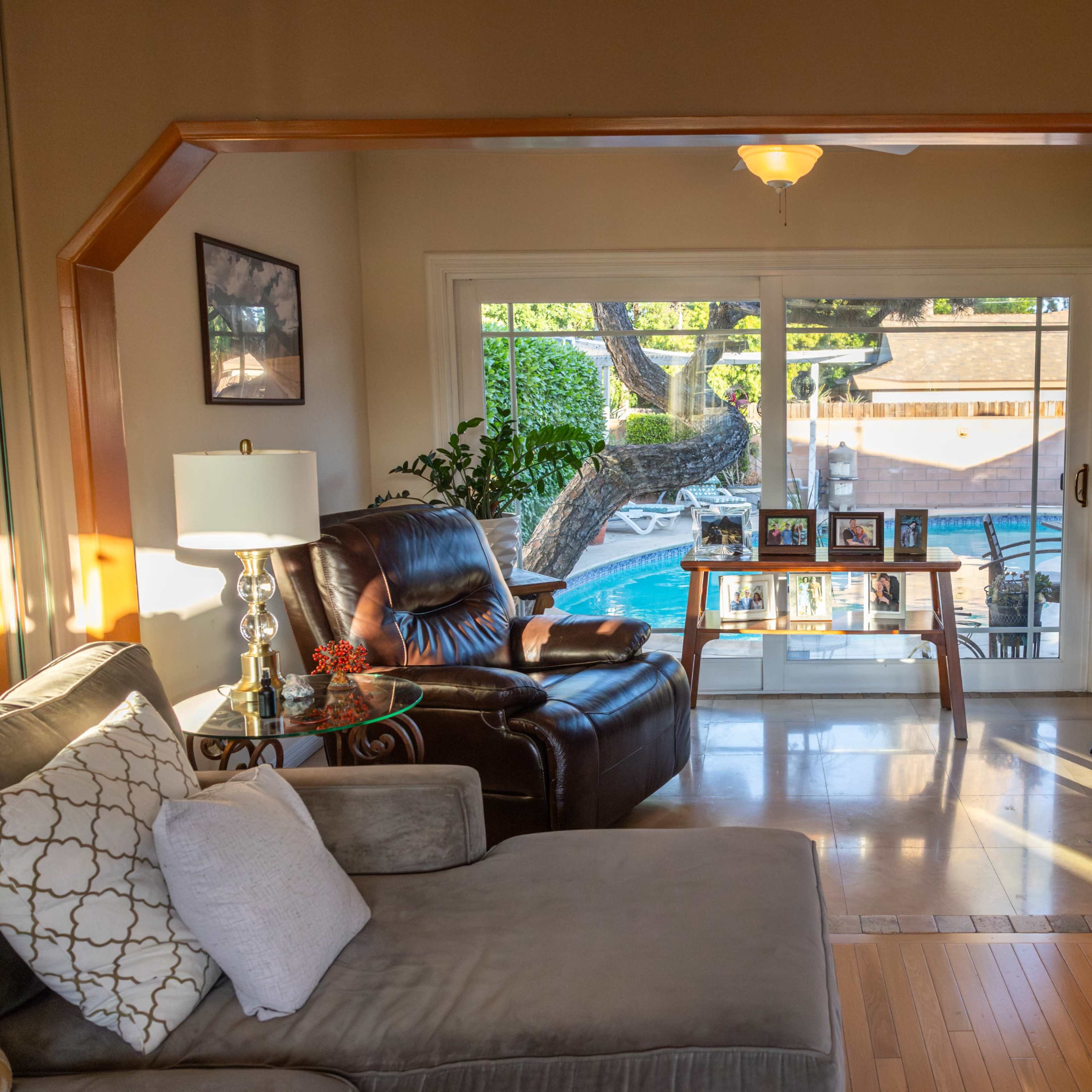 A living room with a view of a backyard pool, featuring a comfortable sofa, an armchair, and a glass table.