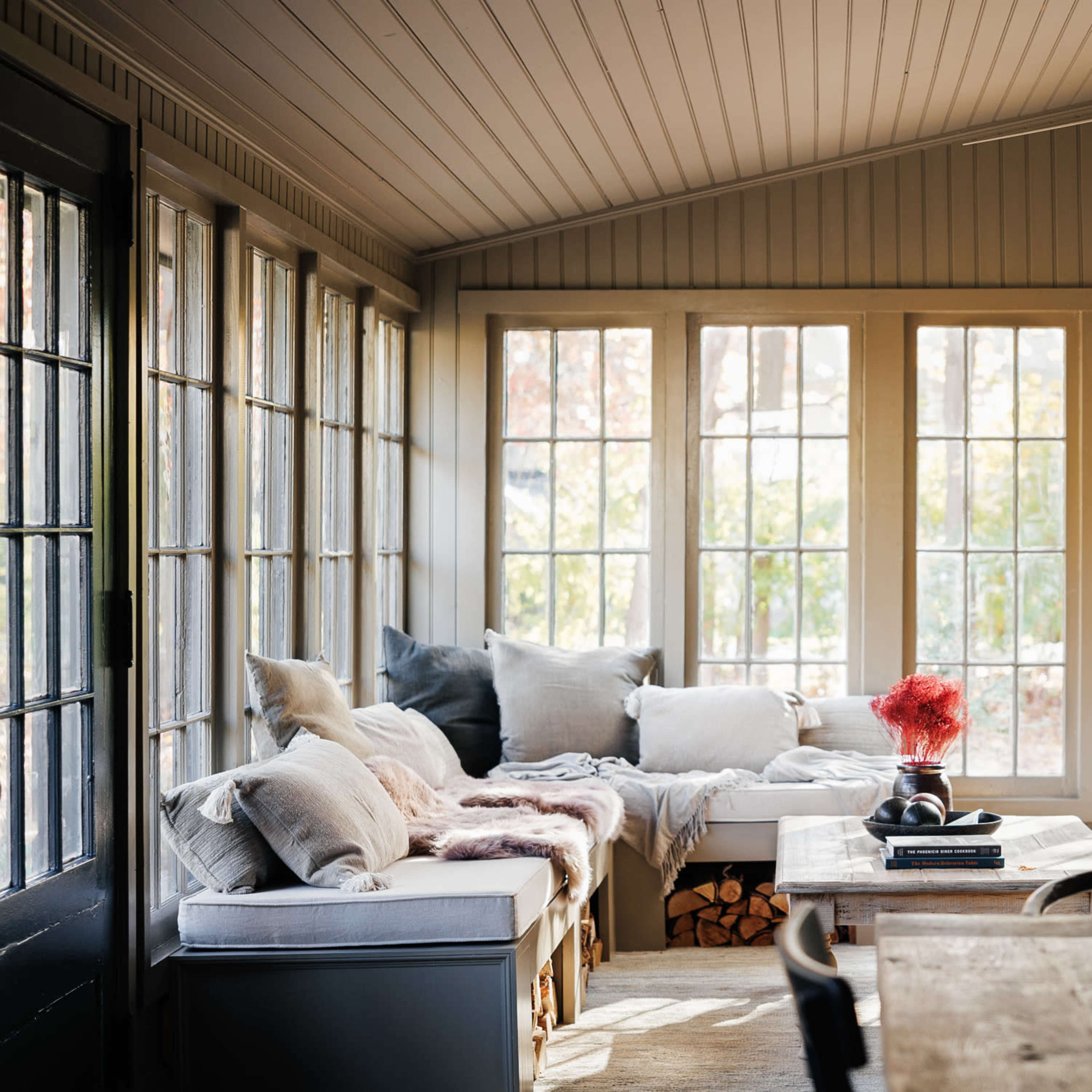 A sunlit room with large windows, a wood stove, a table, and cozy seating areas filled with pillows and blankets.