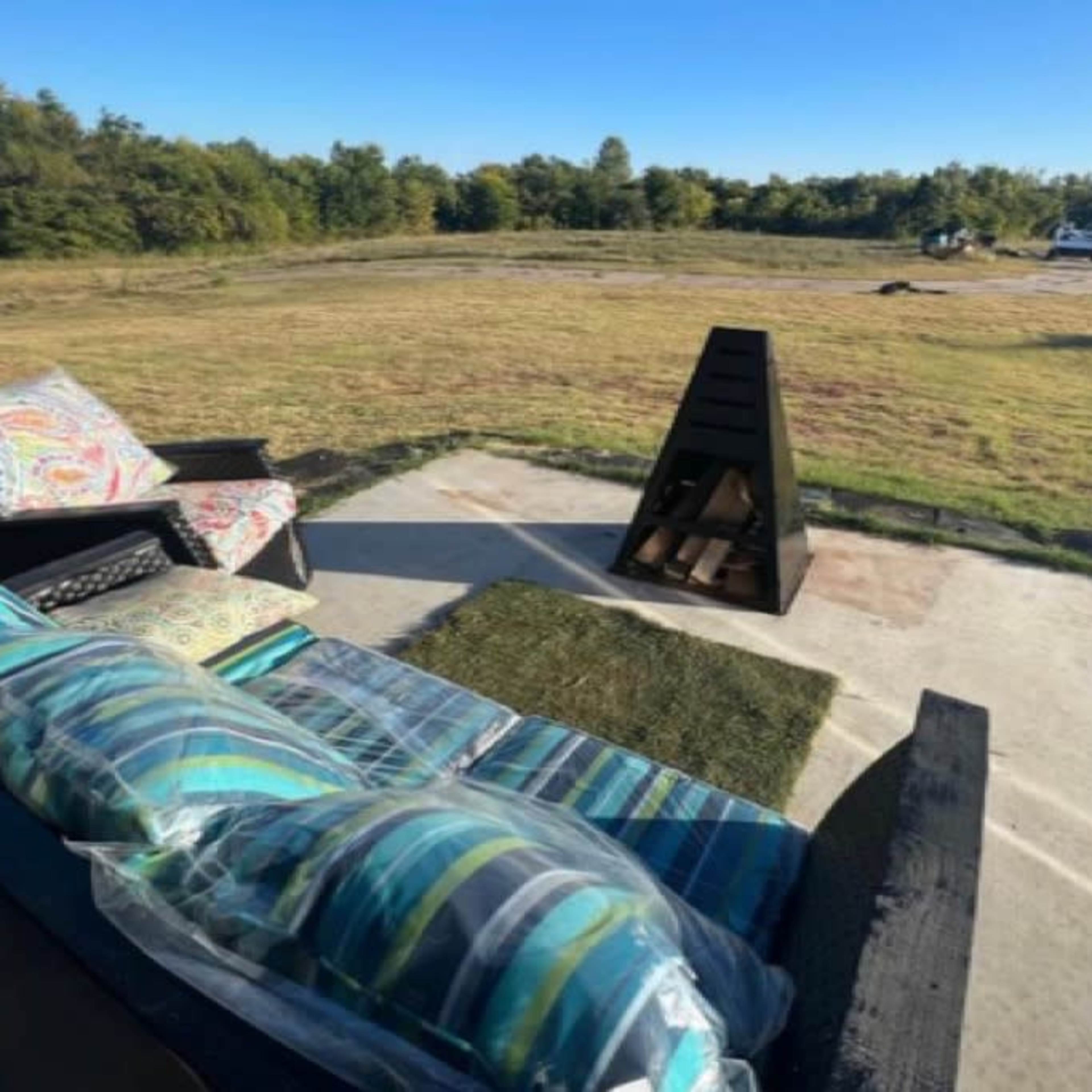 A seating area with striped cushions faces a black outdoor fireplace on a grassy patch, surrounded by trees and an open field.