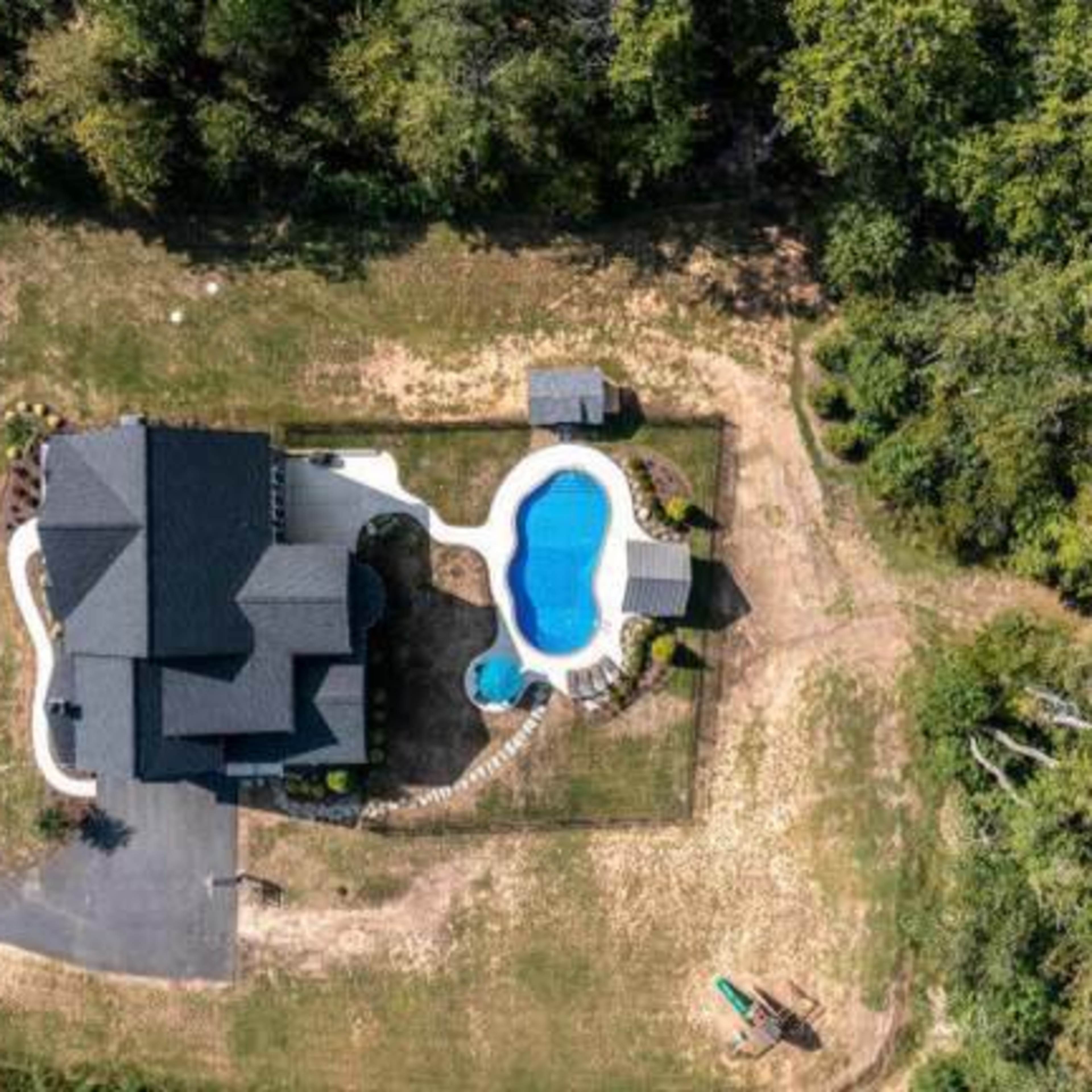 An aerial view of a large house with a swimming pool and surrounding landscape, featuring a driveway and a wooded area nearby.