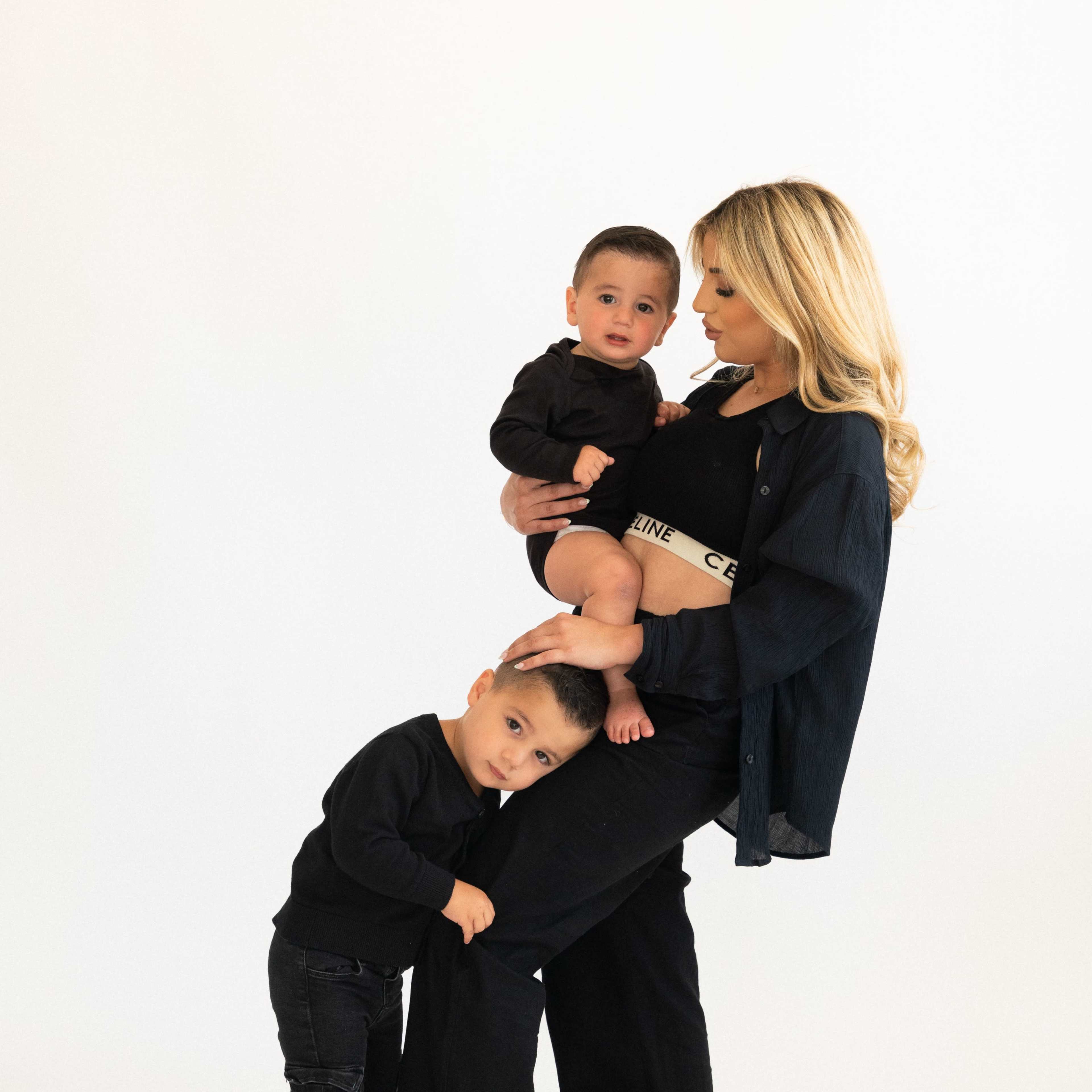 A woman stands with two children, one on her hip and the other holding her leg, all dressed in black against a white background.