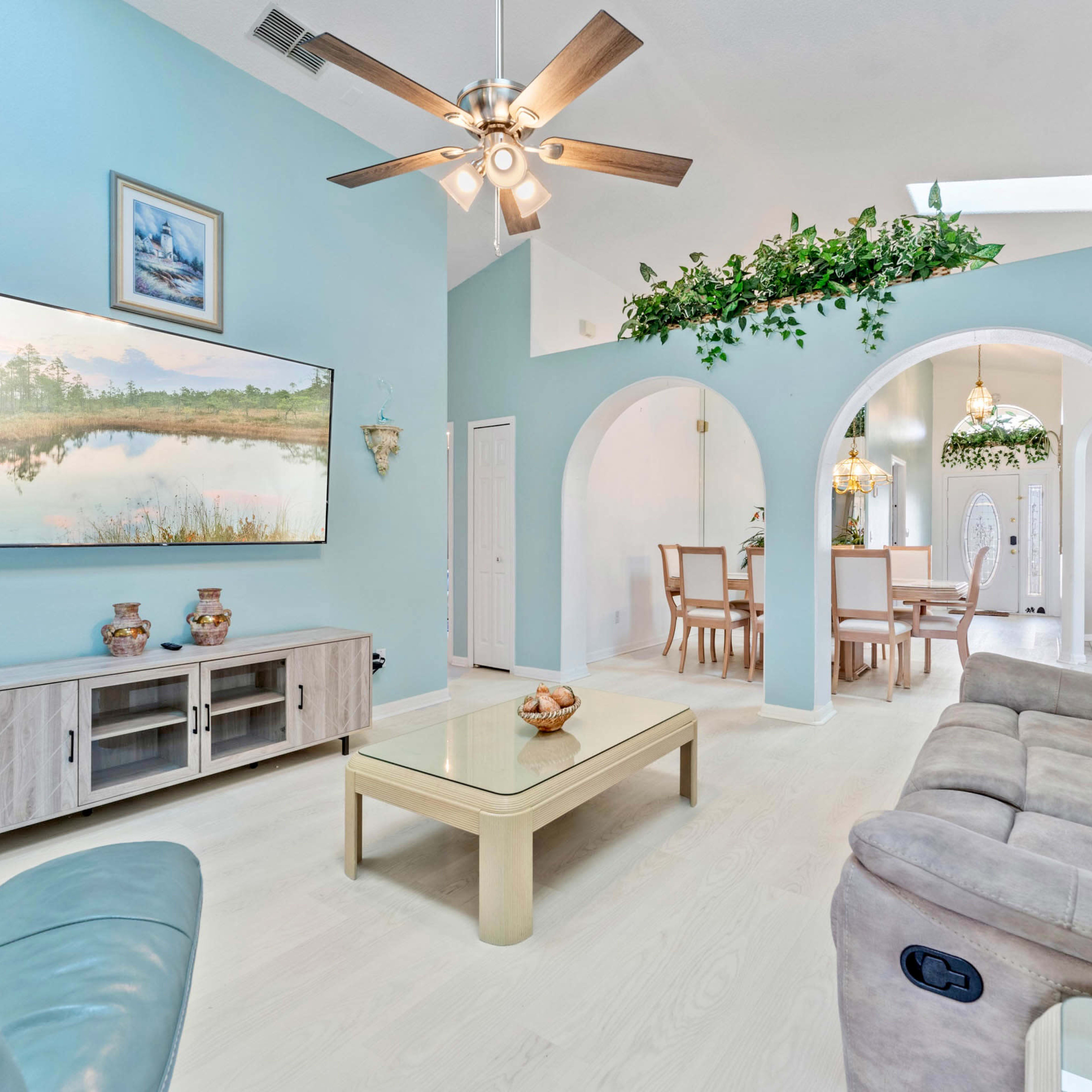 A bright living room with a sectional sofa, a television mounted on the wall, and an adjacent dining area featuring arches and greenery.