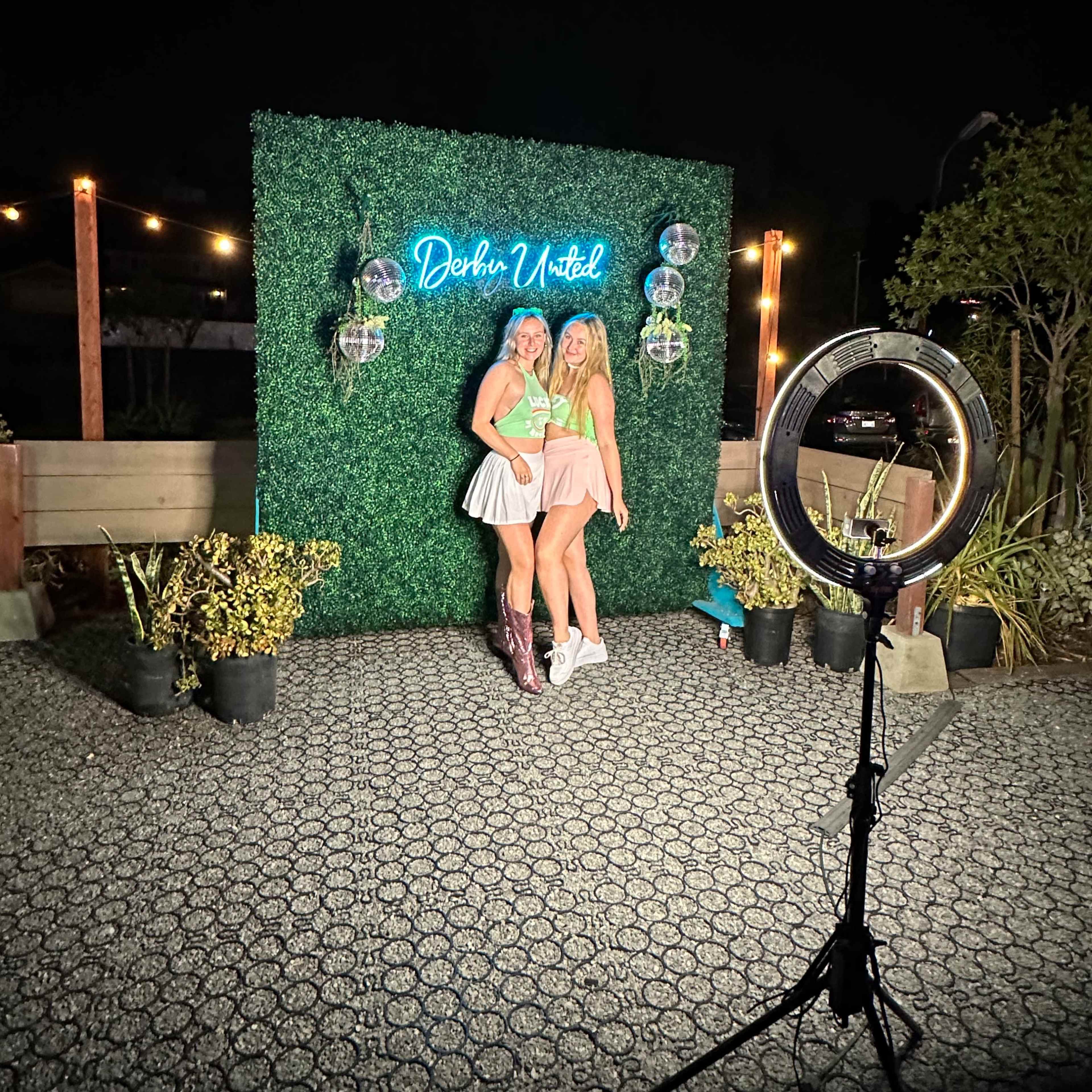 Two women pose in front of a green hedge backdrop with a neon sign that reads "Darlin' Withed," with a ring light illuminating the scene.