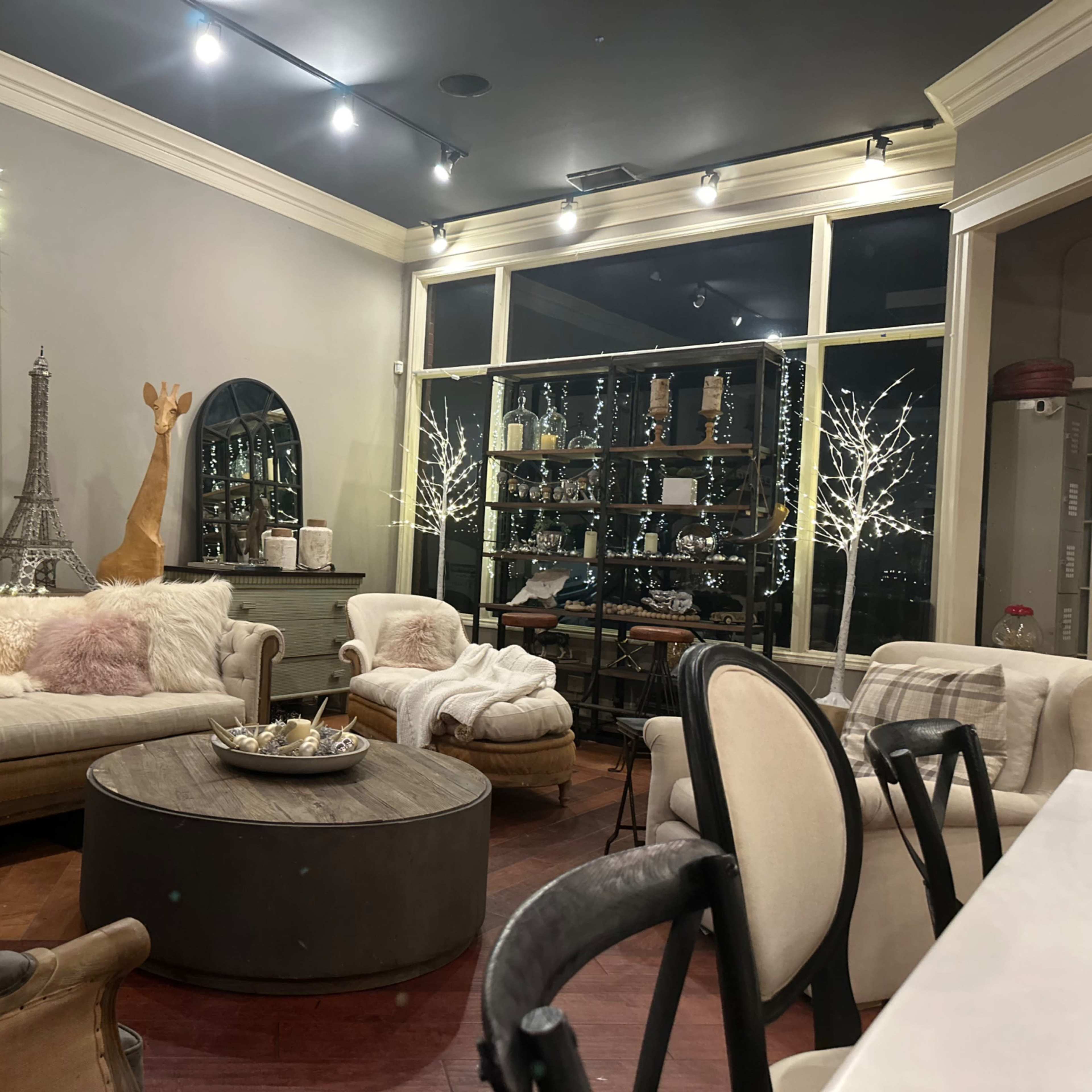 A cozy living space with a round coffee table, plush sofas, decorative mirrors, and a large window displaying twinkling lights outside.