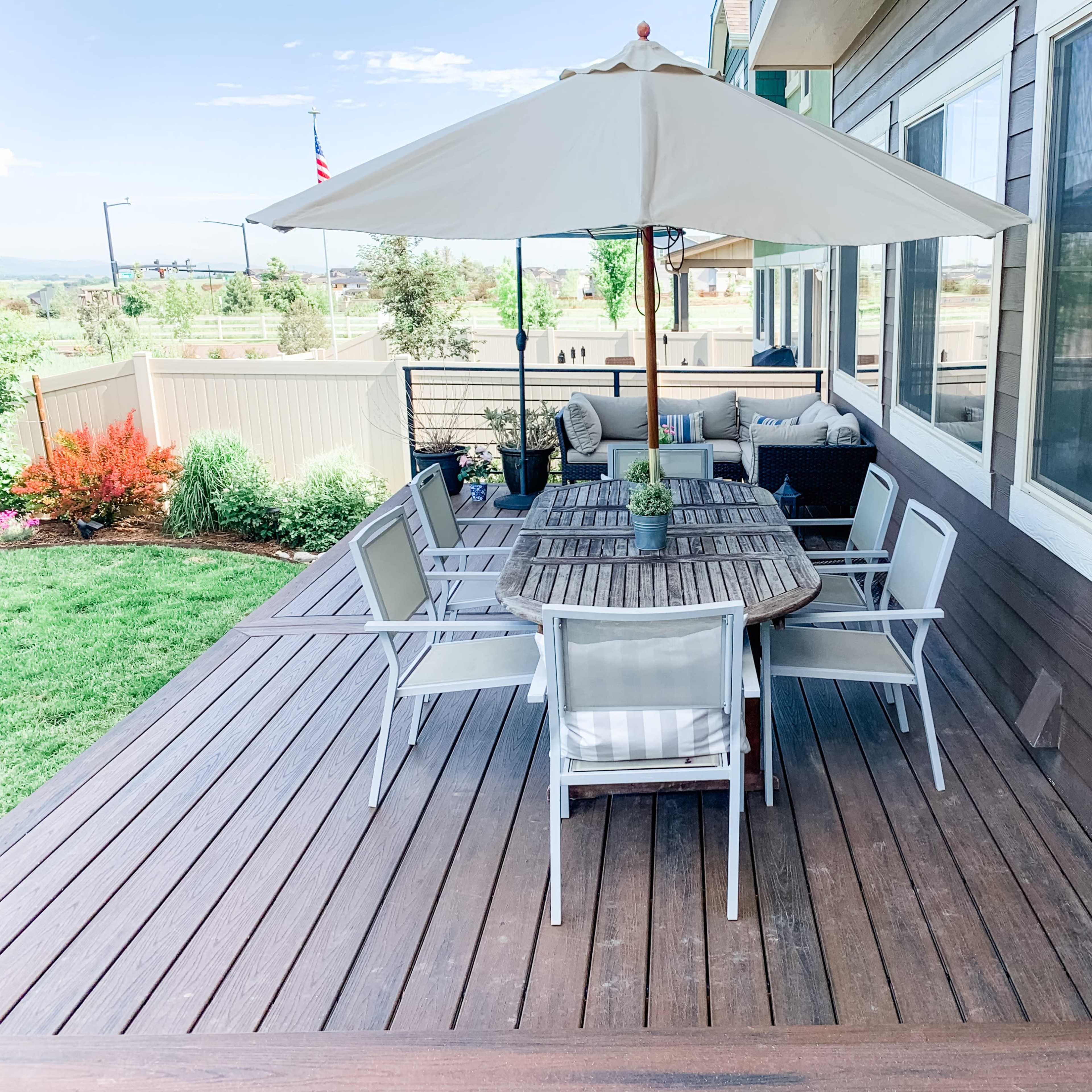 A wooden deck with a dining table, six chairs, and an umbrella, surrounded by landscaped grass and plants.