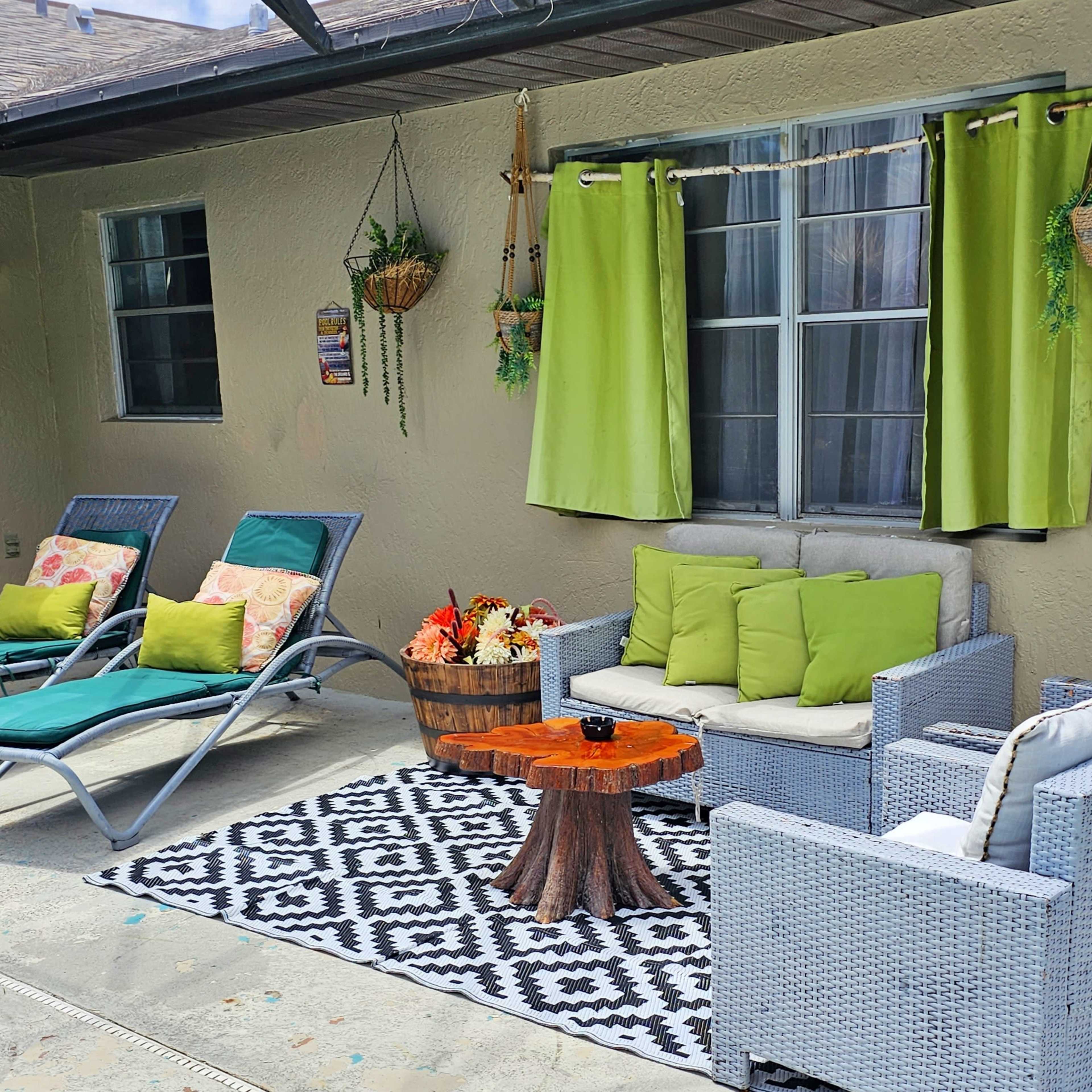 A patio area featuring two lounge chairs, a seating arrangement with green cushions, and a wooden table on a patterned rug.