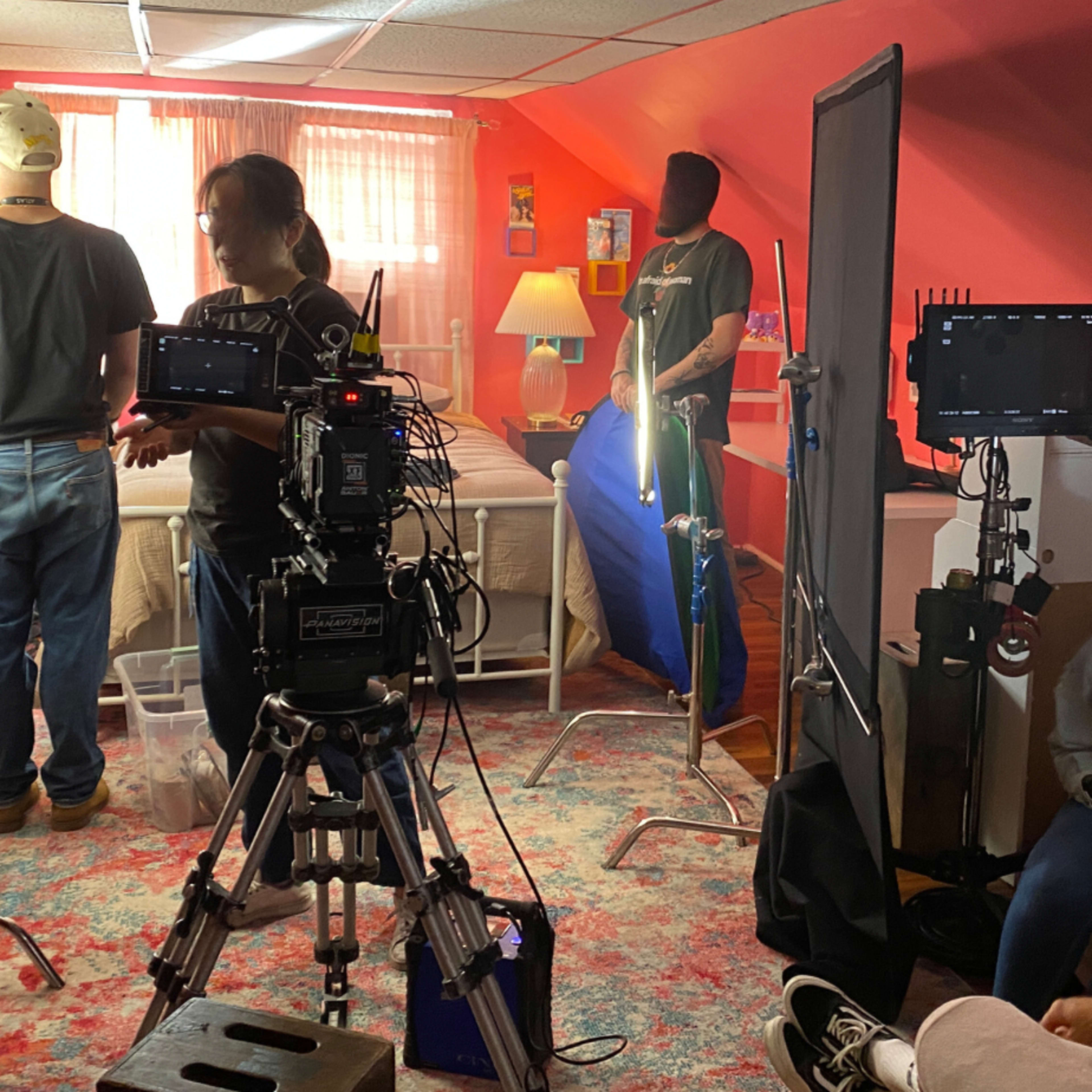 A film crew sets up lighting and equipment in a room with a bed and colorful decor for a shoot.
