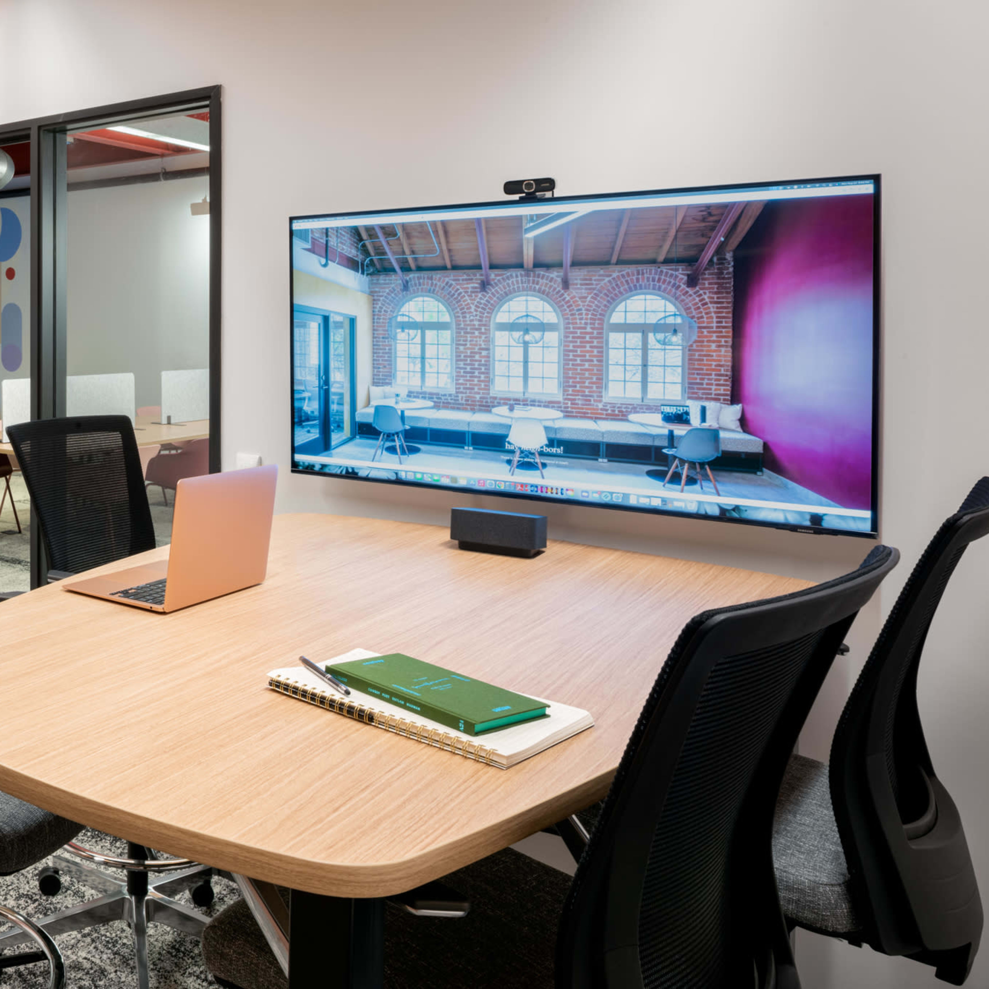 A modern meeting room features a wooden table with a laptop and a large screen displaying a vibrant image of a workspace, surrounded by ergonomic chairs.