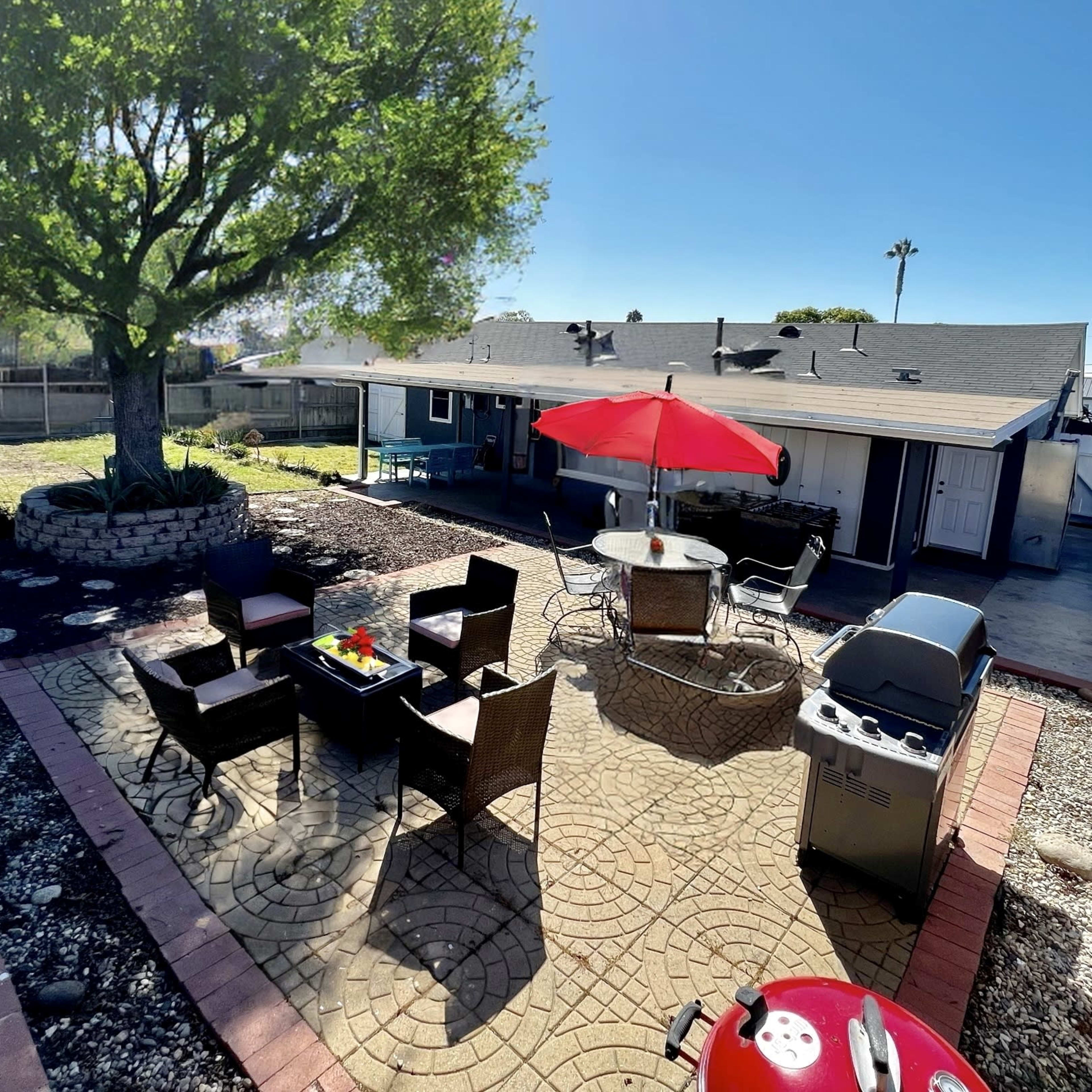 A backyard patio area with a stone pathway, outdoor seating arrangement, a grill, and a large tree providing shade.