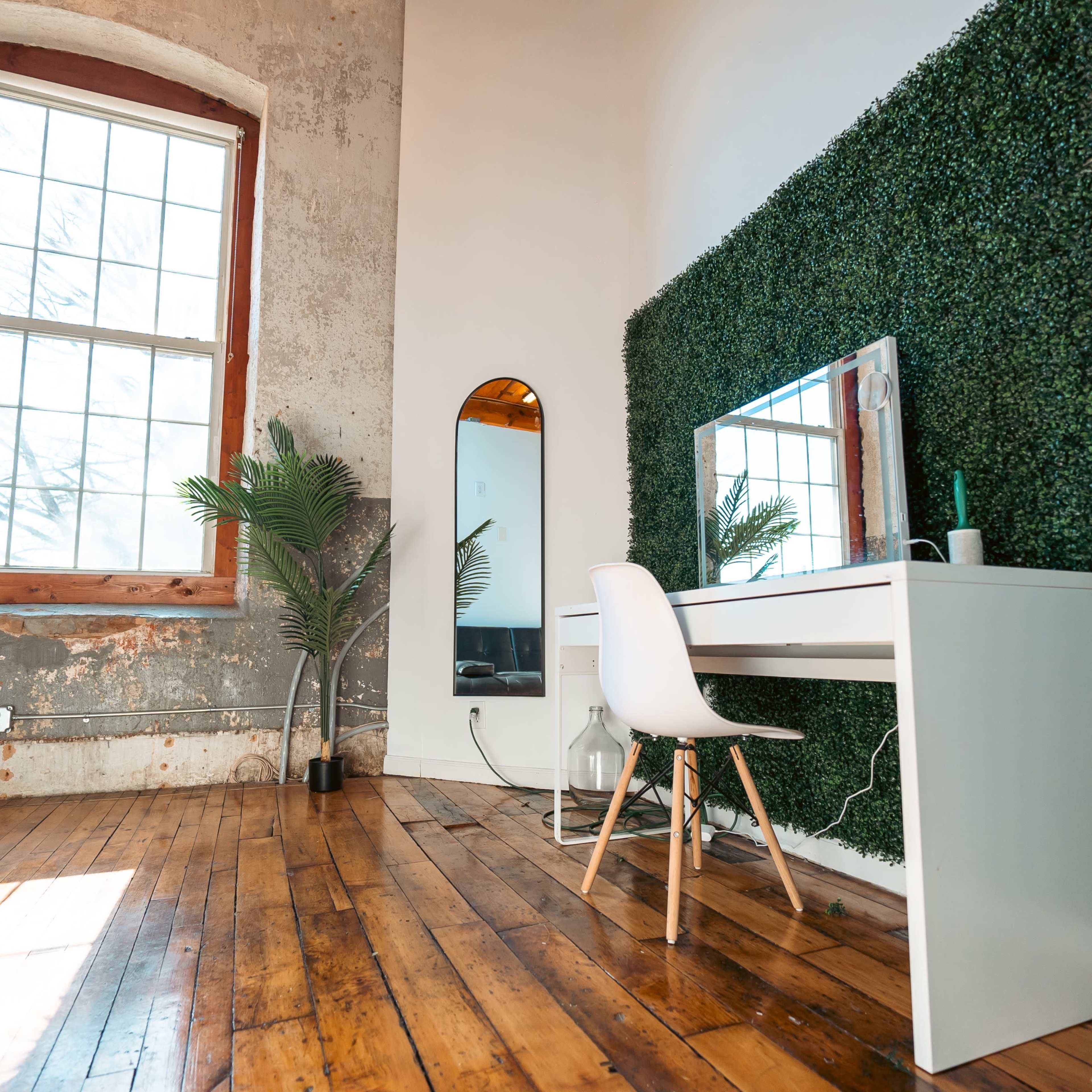 A modern workspace with a white desk and chair against a green wall of artificial foliage, featuring a large window that lets in natural light.