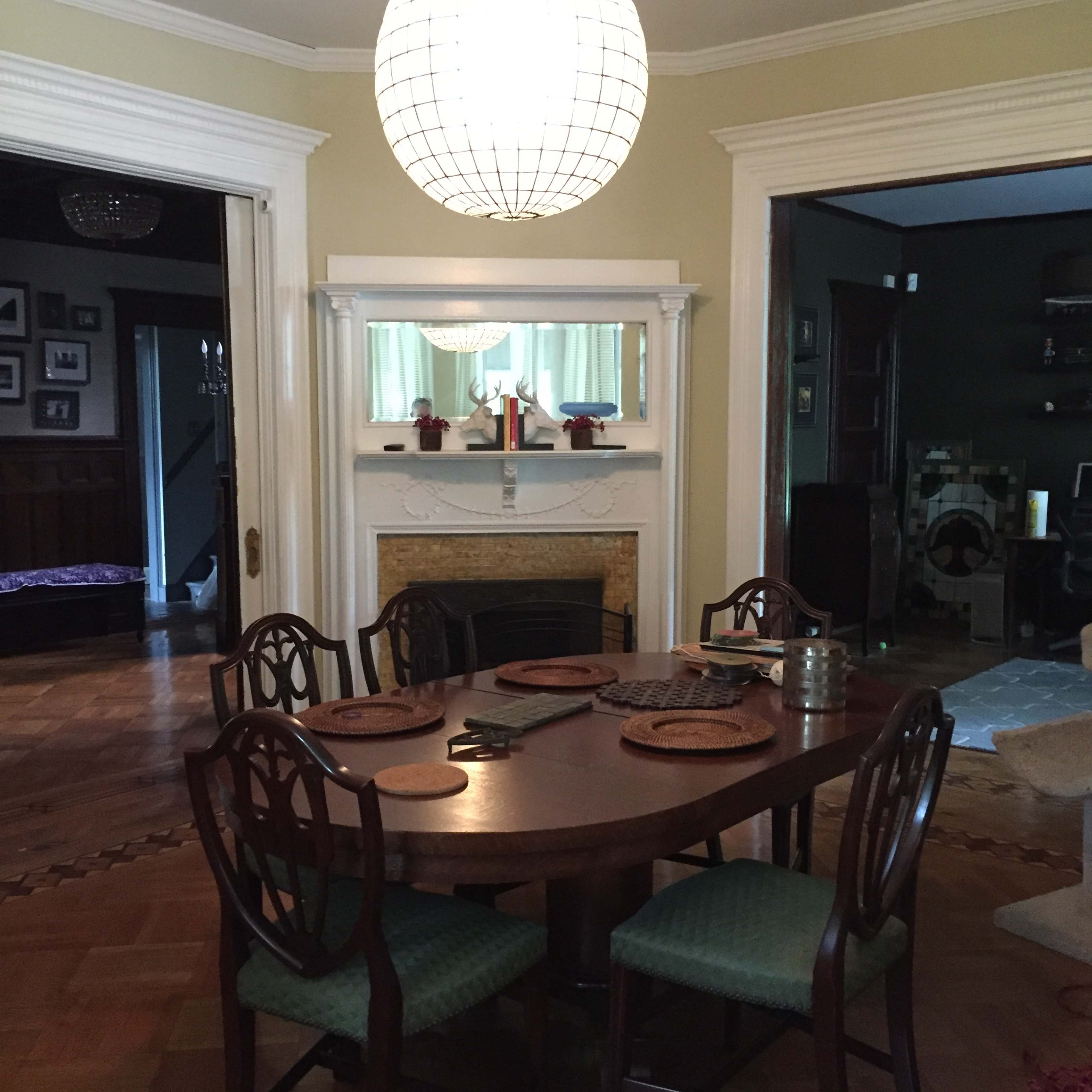 A round wooden dining table is set in a well-lit room, surrounded by four chairs, beneath a large pendant light.
