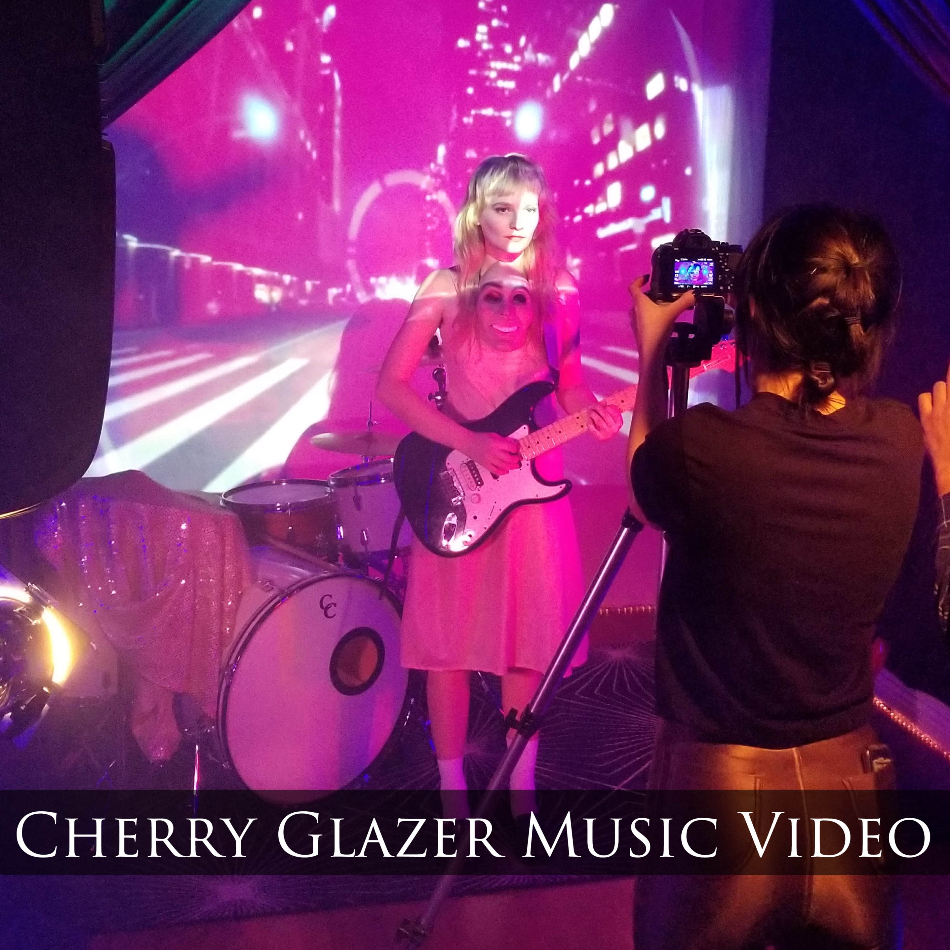 A musician performs on stage in front of a projected cityscape while being filmed by a camera operator.