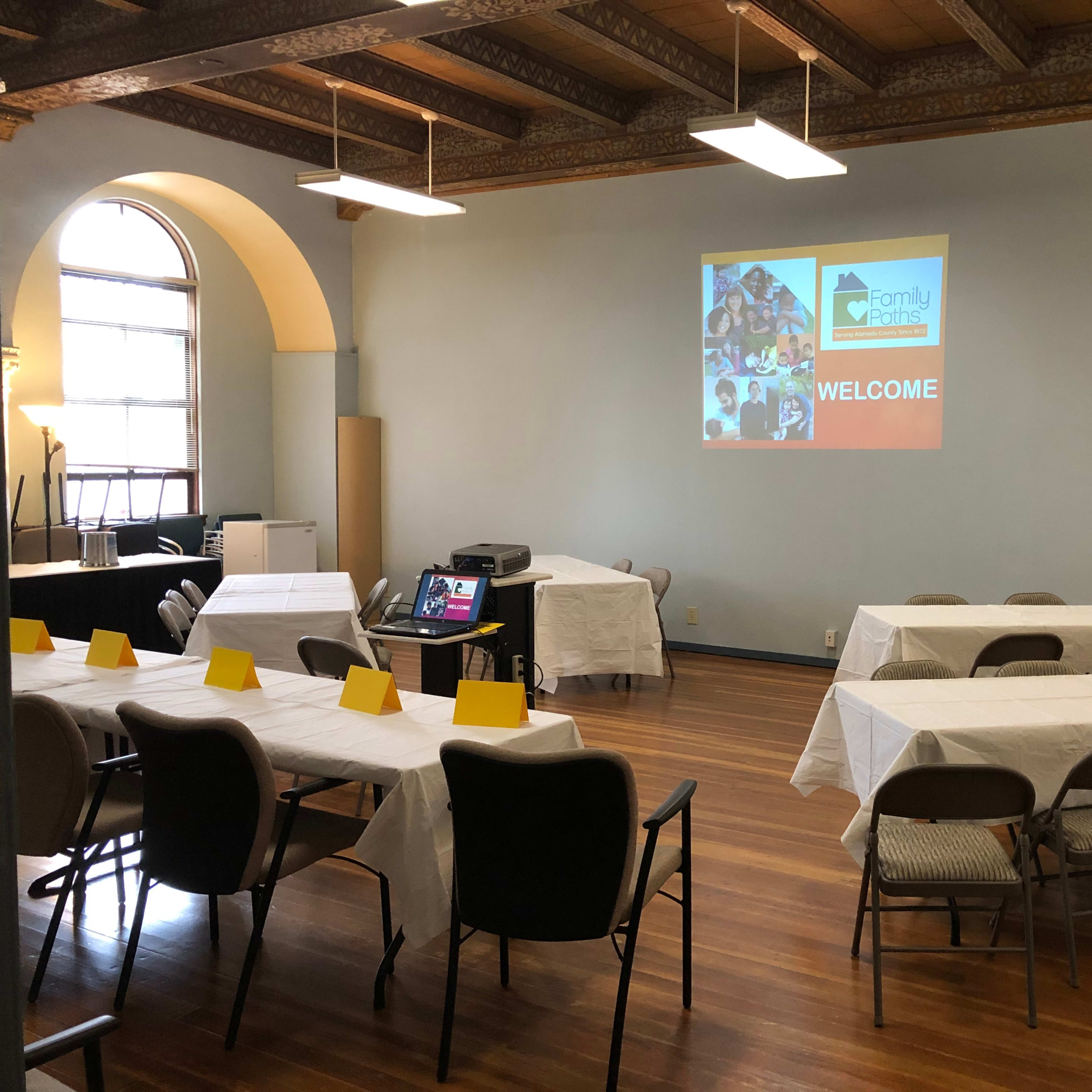 A conference room setup with several tables covered in white tablecloths, chairs arranged around them, and a projector displaying a "WELCOME" message on the wall.