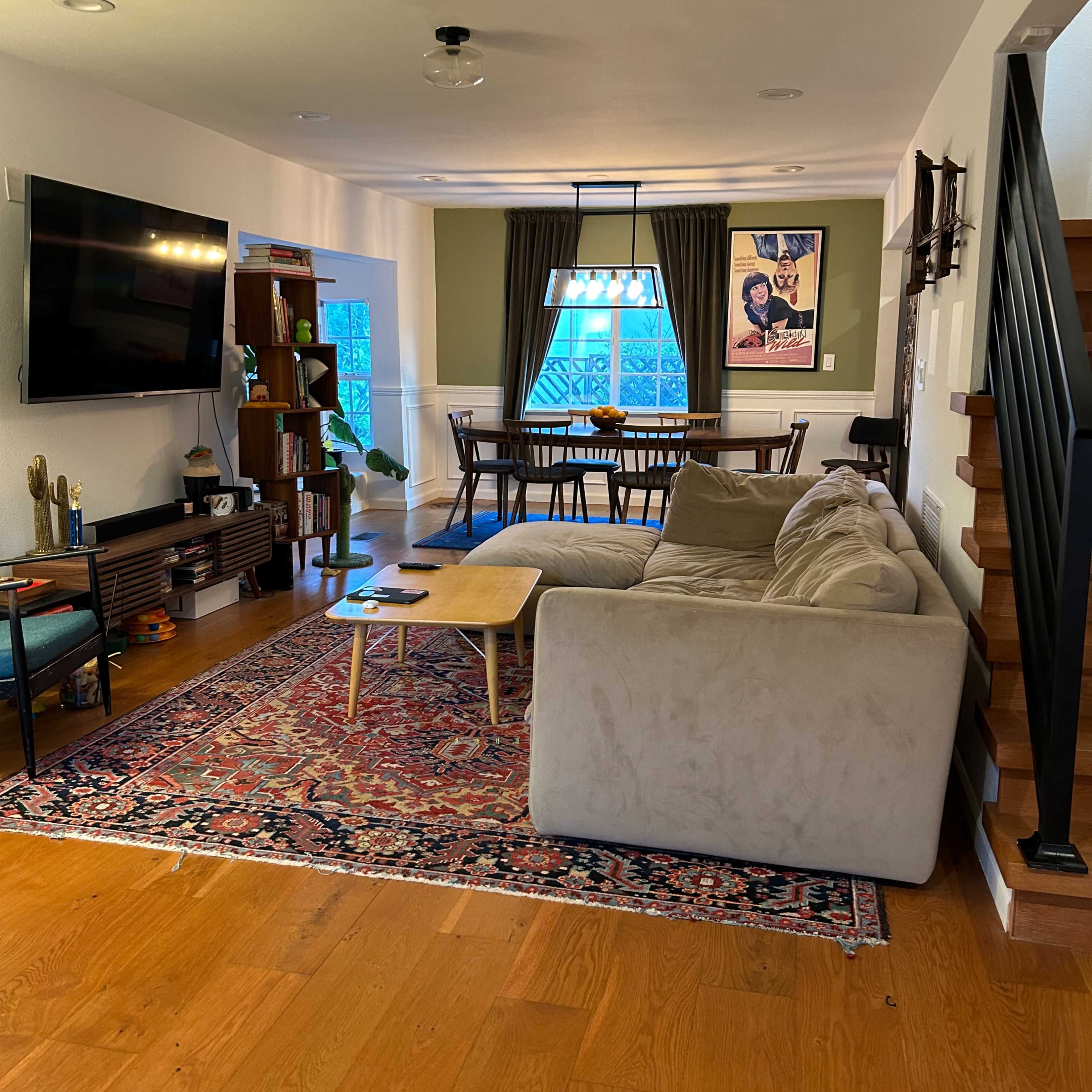 A spacious living room with a beige sectional sofa, a large television mounted on the wall, and a wooden dining table set against green walls.