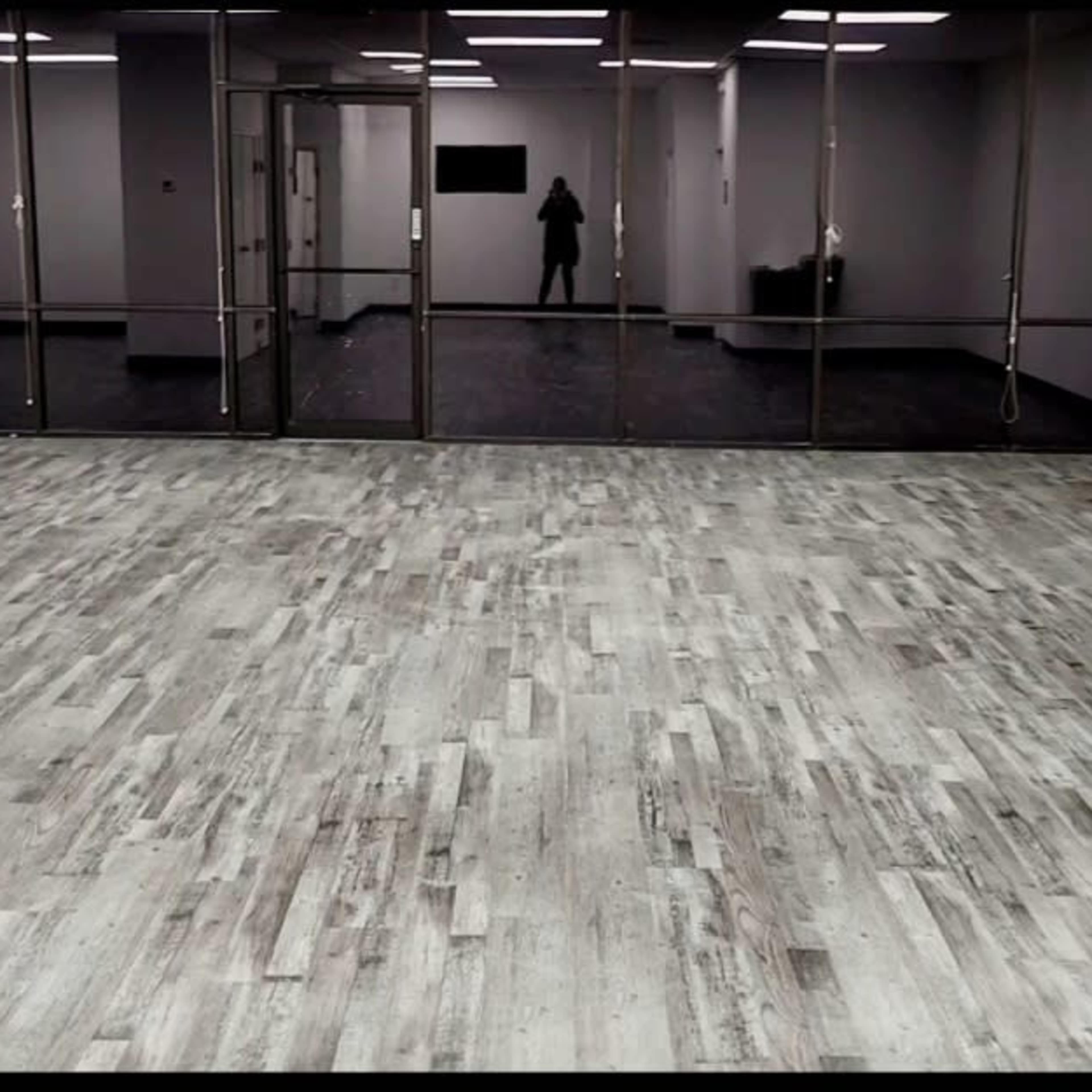 An empty room with a wooden floor and large glass windows reflecting a figure in the background.