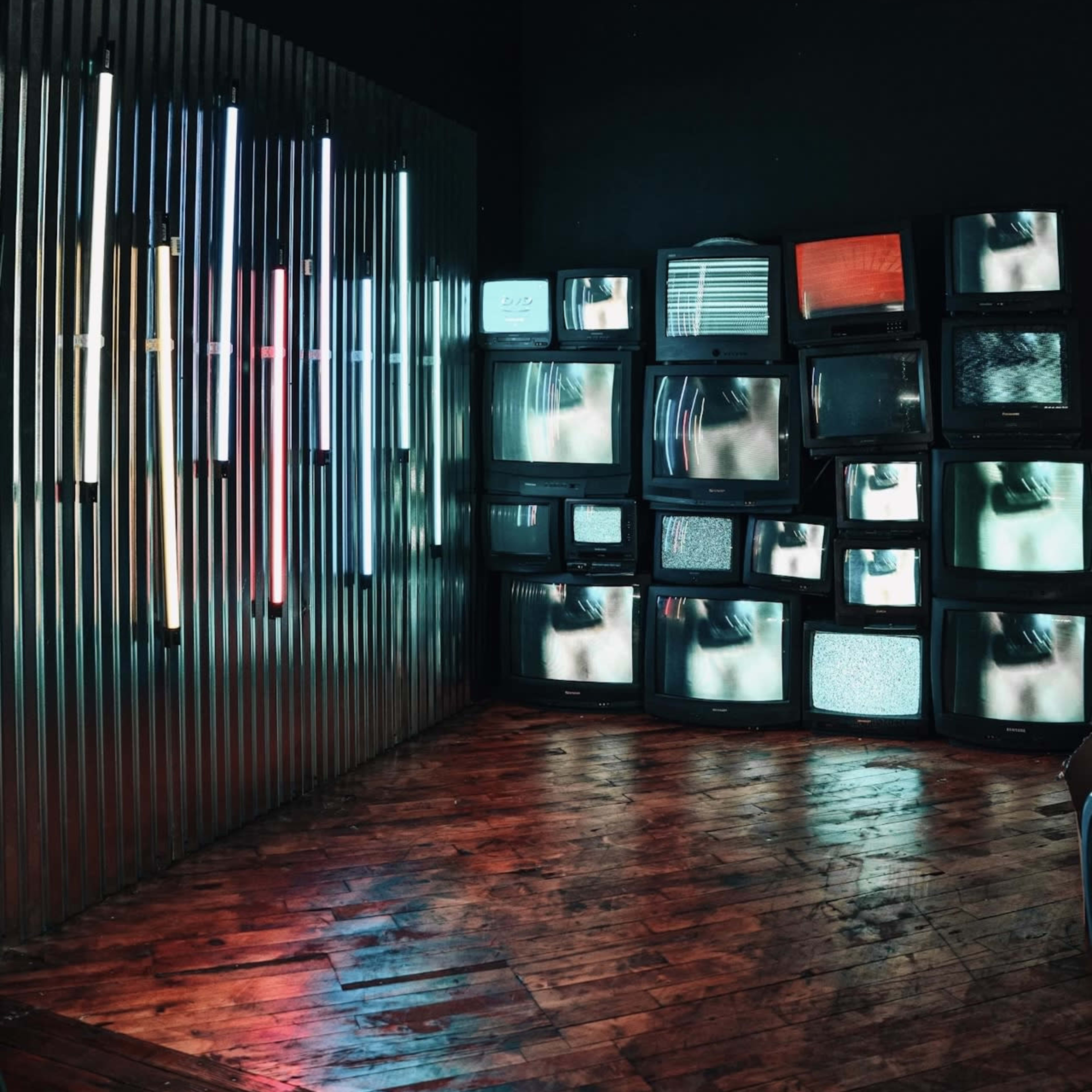 A corner of a room features multiple vintage televisions displaying various static images, alongside a wall of illuminated neon lights.