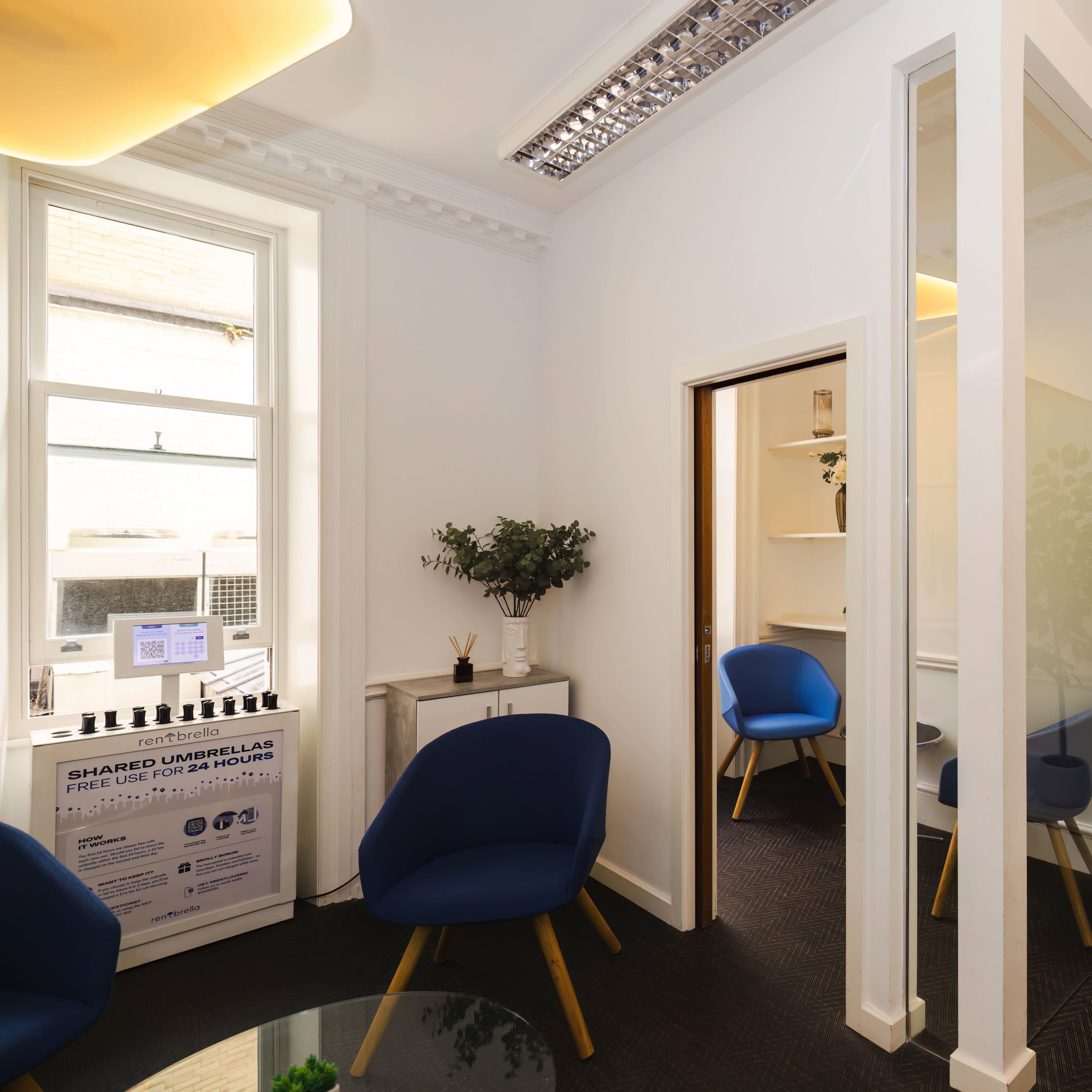A modern office space with blue chairs, a glass partition, a wall clock, and a small shelf with plants.