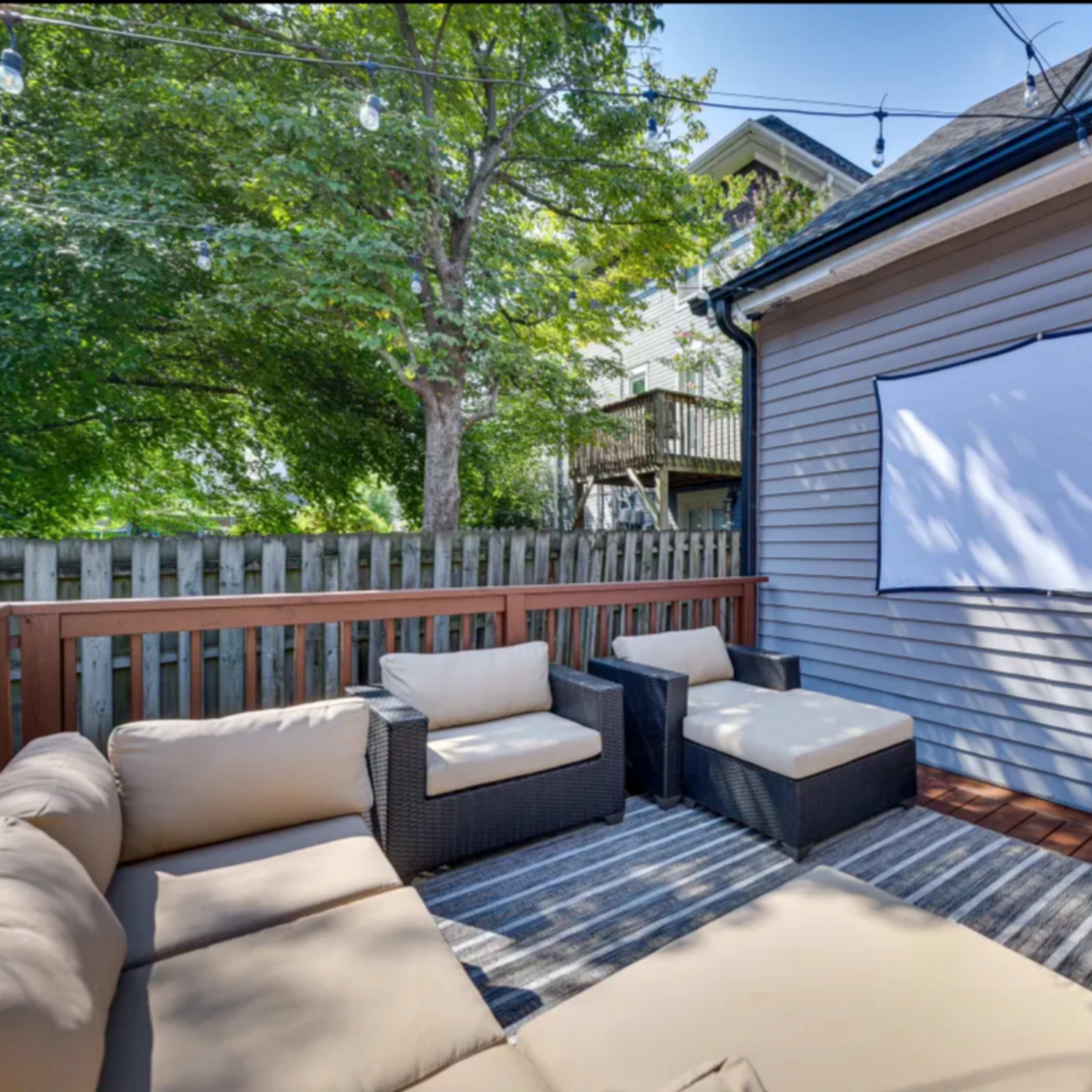 A patio area with a seating arrangement of wicker chairs and a light-colored couch, facing a blank projection screen mounted on a wooden fence.