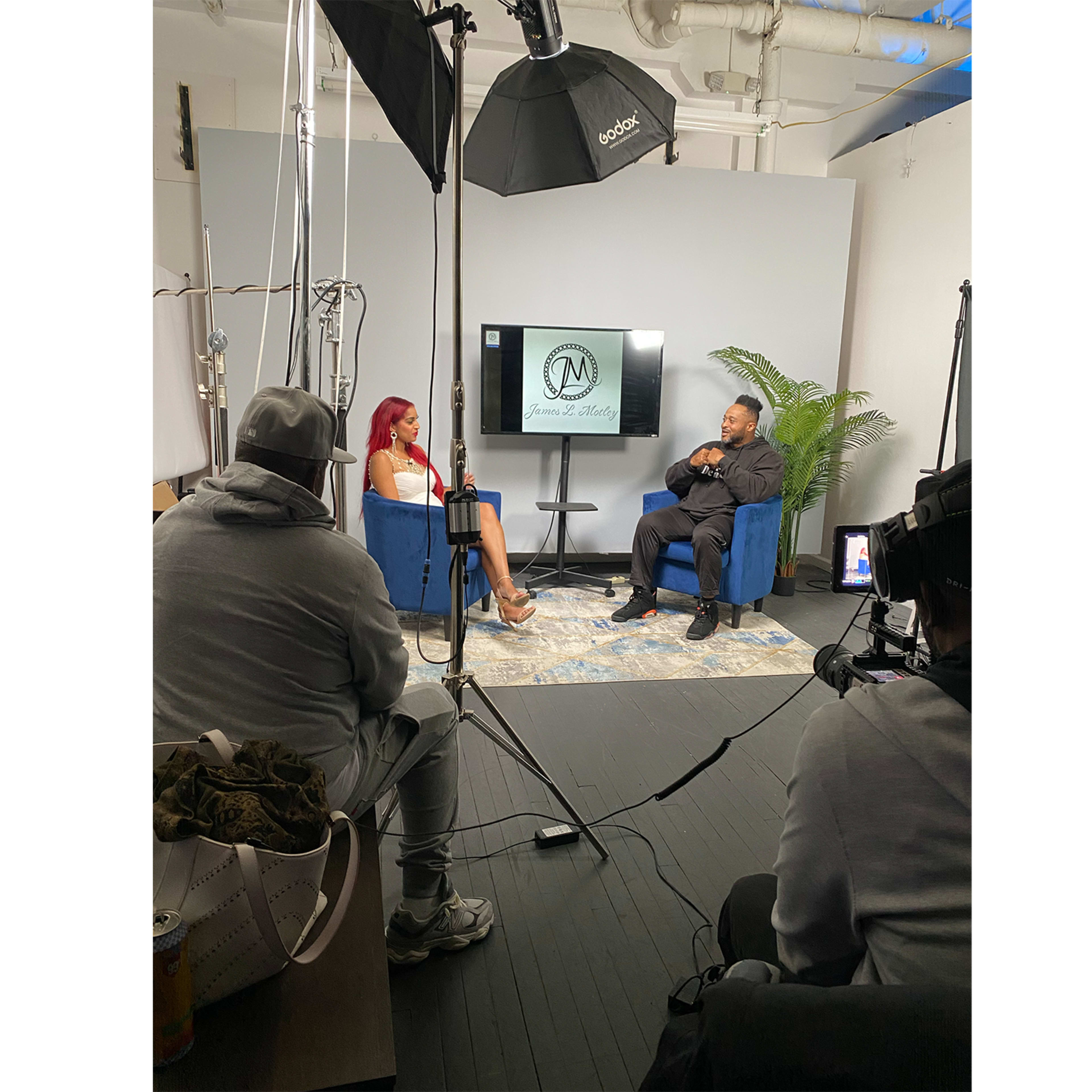 A woman and a man are seated in blue chairs during an interview in a studio setting with a camera crew and lighting equipment.
