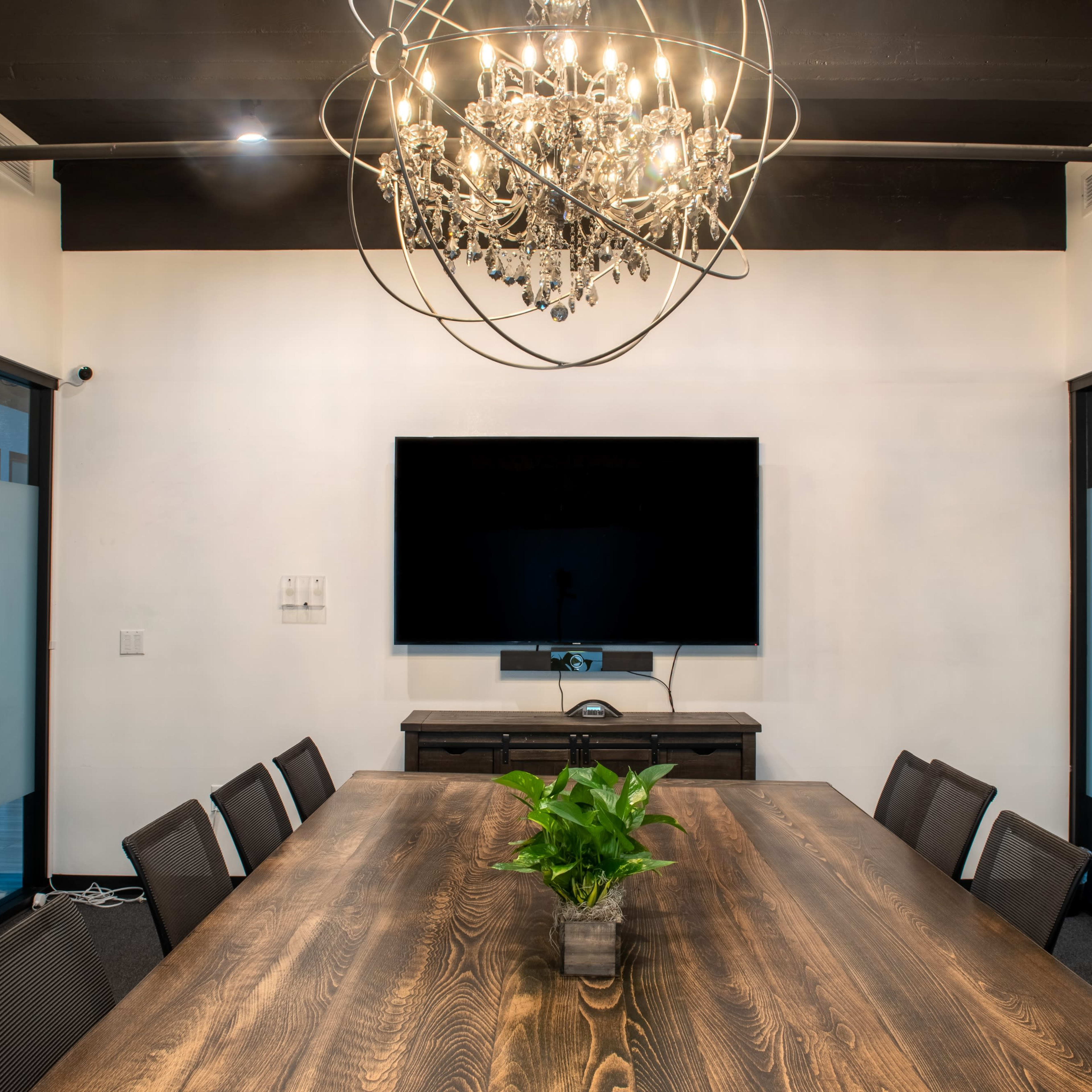 A long wooden conference table is set in a modern meeting room with a chandelier above and a large TV mounted on the wall.