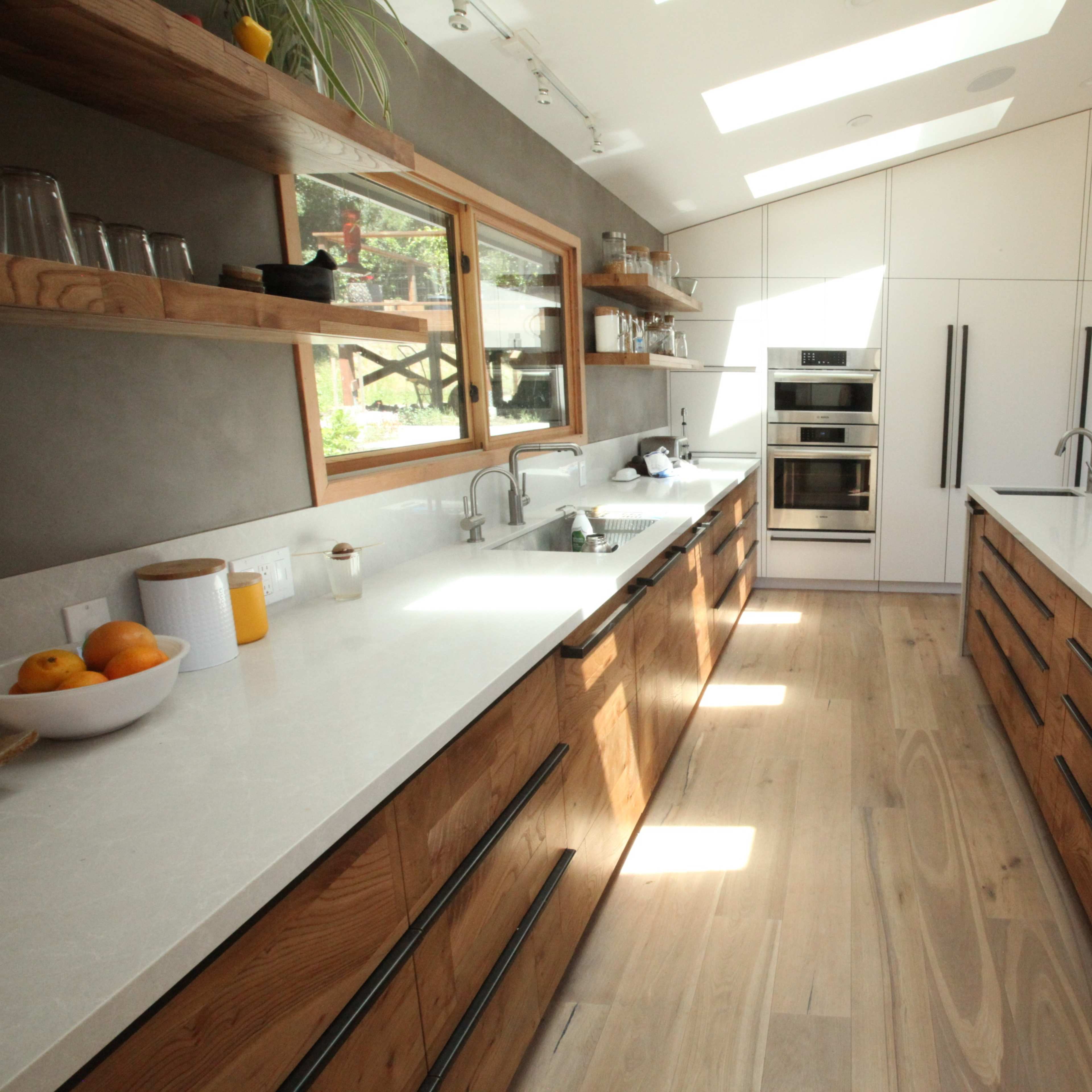 A modern kitchen features wooden cabinetry, white countertops, and large windows allowing natural light.