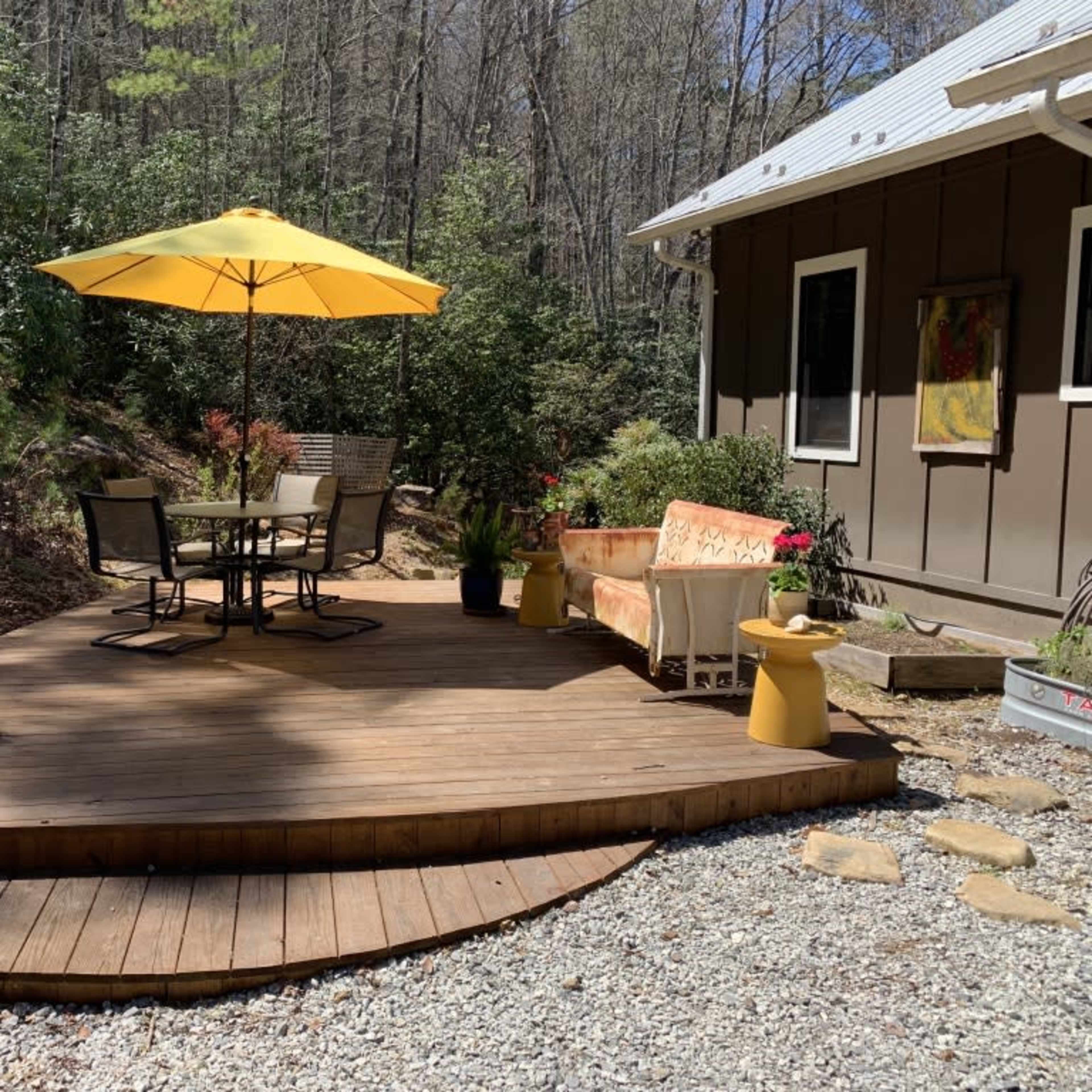 A wooden deck with a round table and four chairs, an umbrella, and a colorful seating area is surrounded by greenery and gravel.