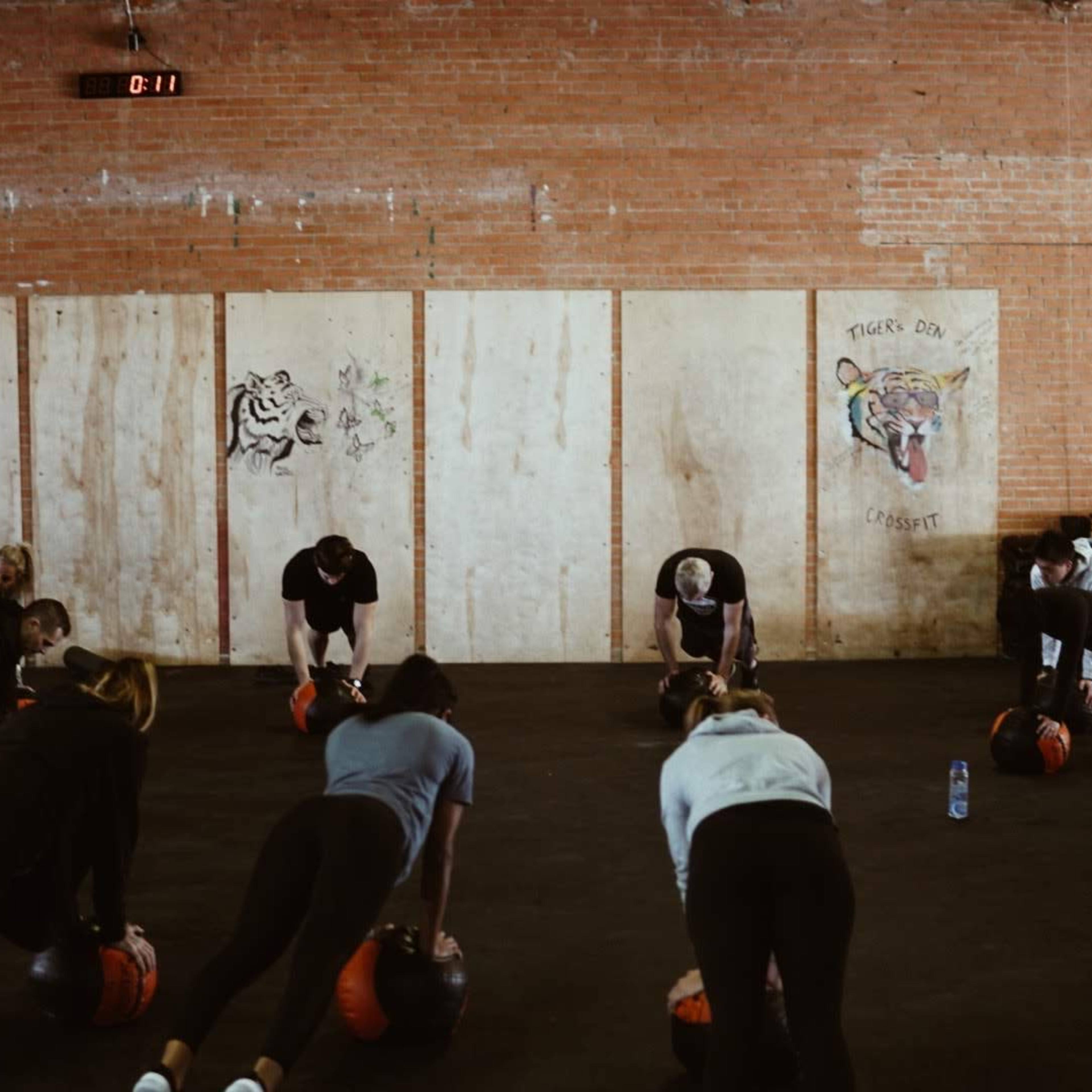 A group of people is performing a workout on a gym floor, using weighted balls while facing down in a plank position.
