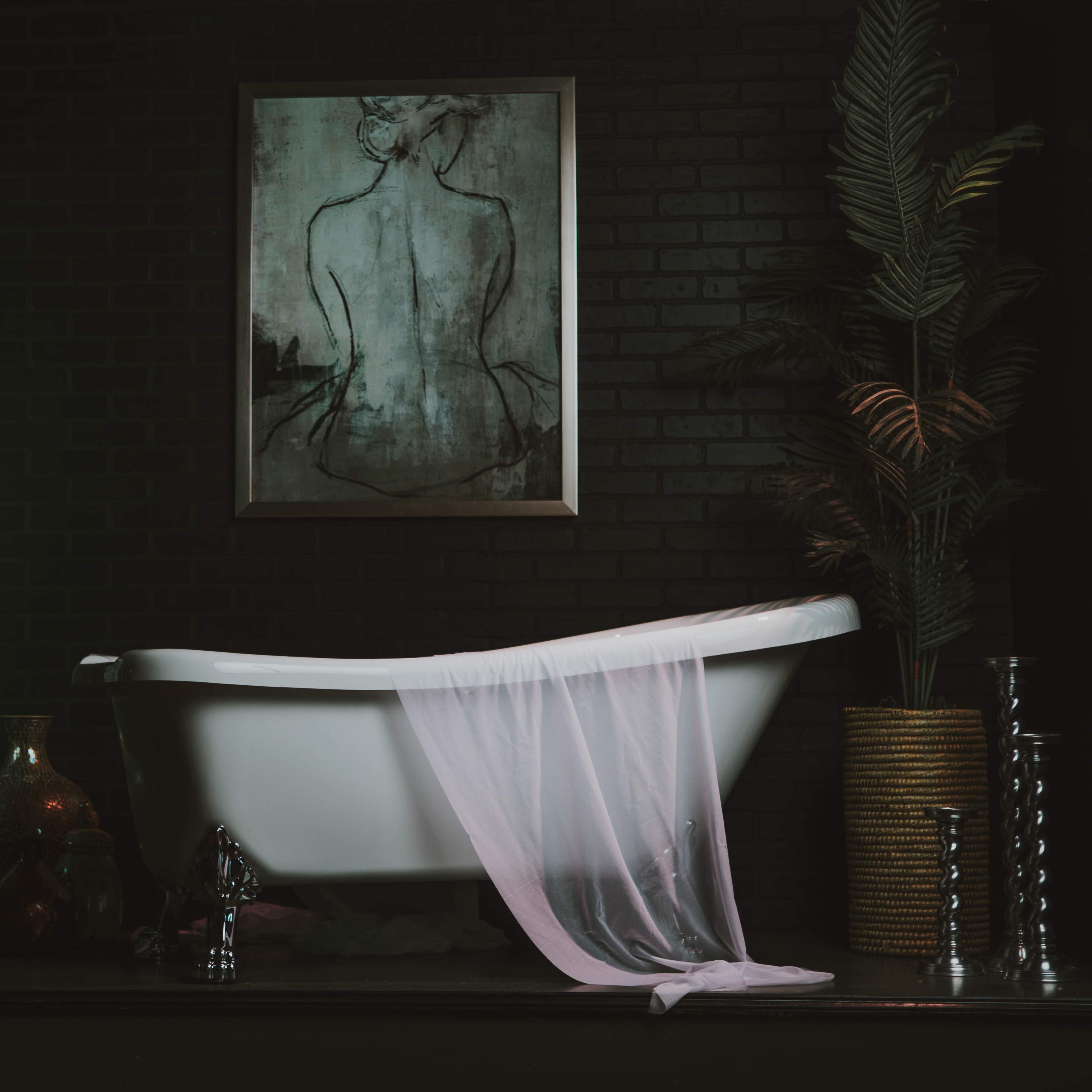 A white freestanding bathtub is positioned against a black brick wall, adorned with a painting and surrounded by plants and decorative vases.