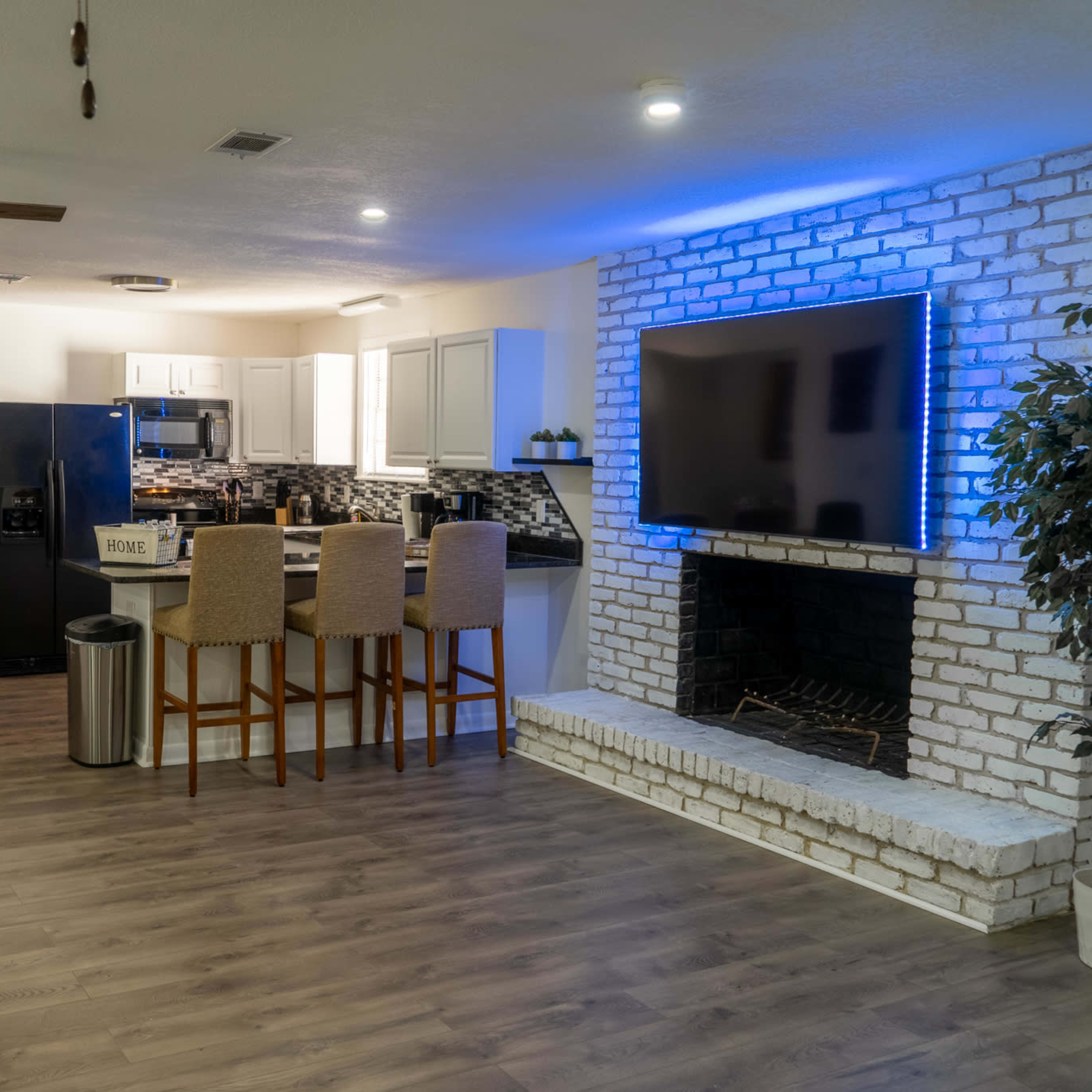 A modern living area with a kitchen and a seating area, featuring a fireplace and a large television mounted on a white brick wall.