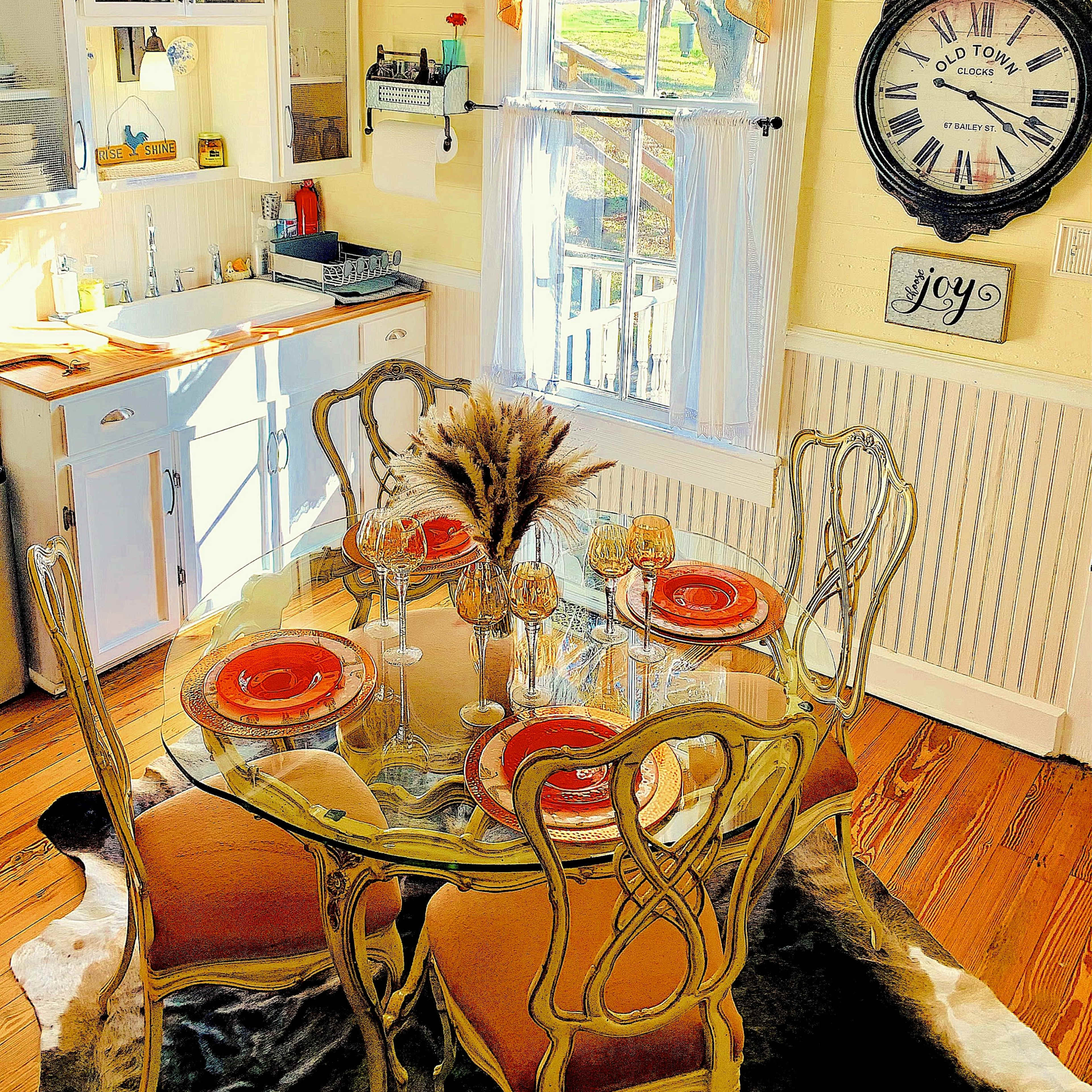 A dining area features a glass table with orange plates and decorative glassware, surrounded by elegant chairs, with a window and kitchen cabinetry in the background.