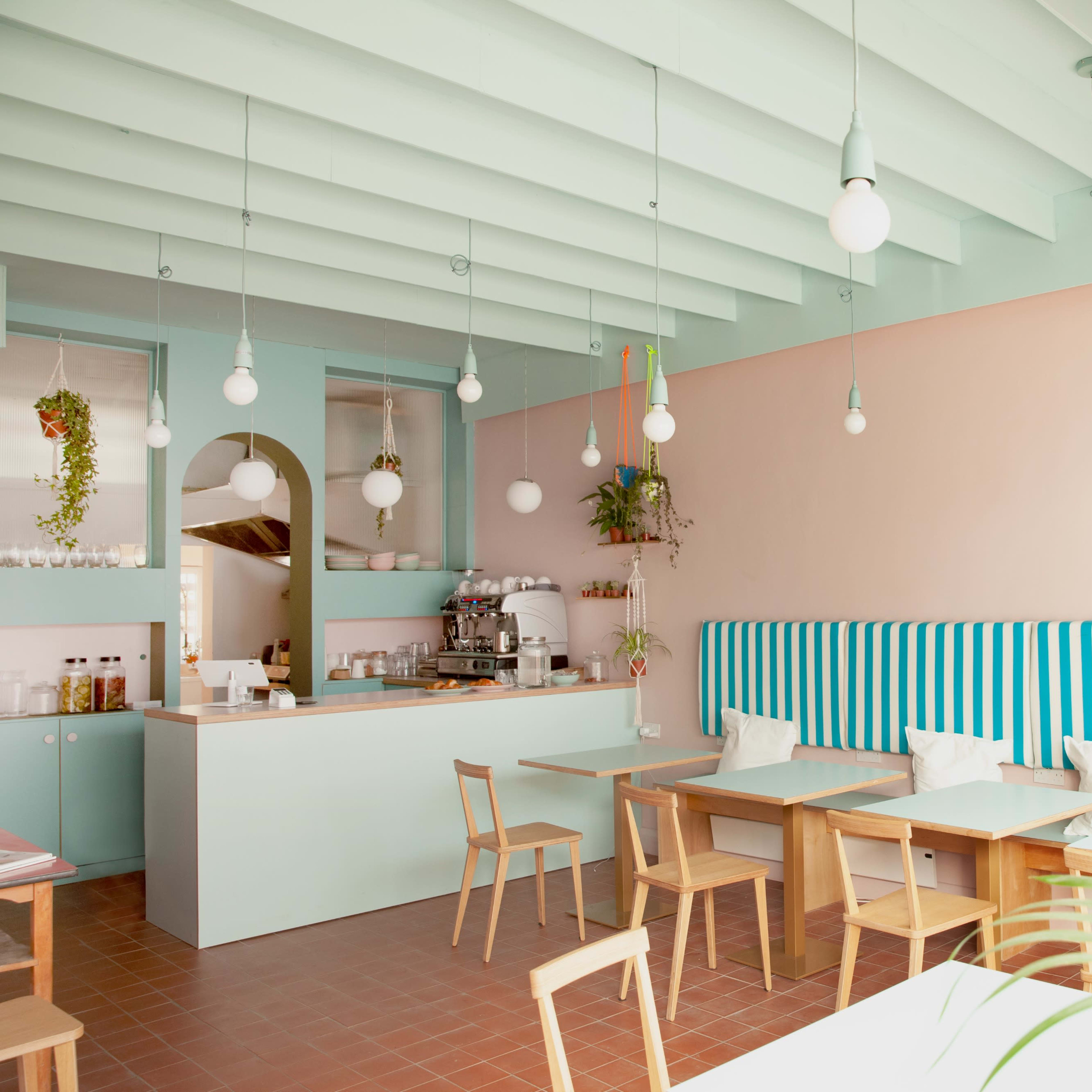 A modern cafe features light blue and pink walls, wooden tables, and striped seating with hanging pendant lights.