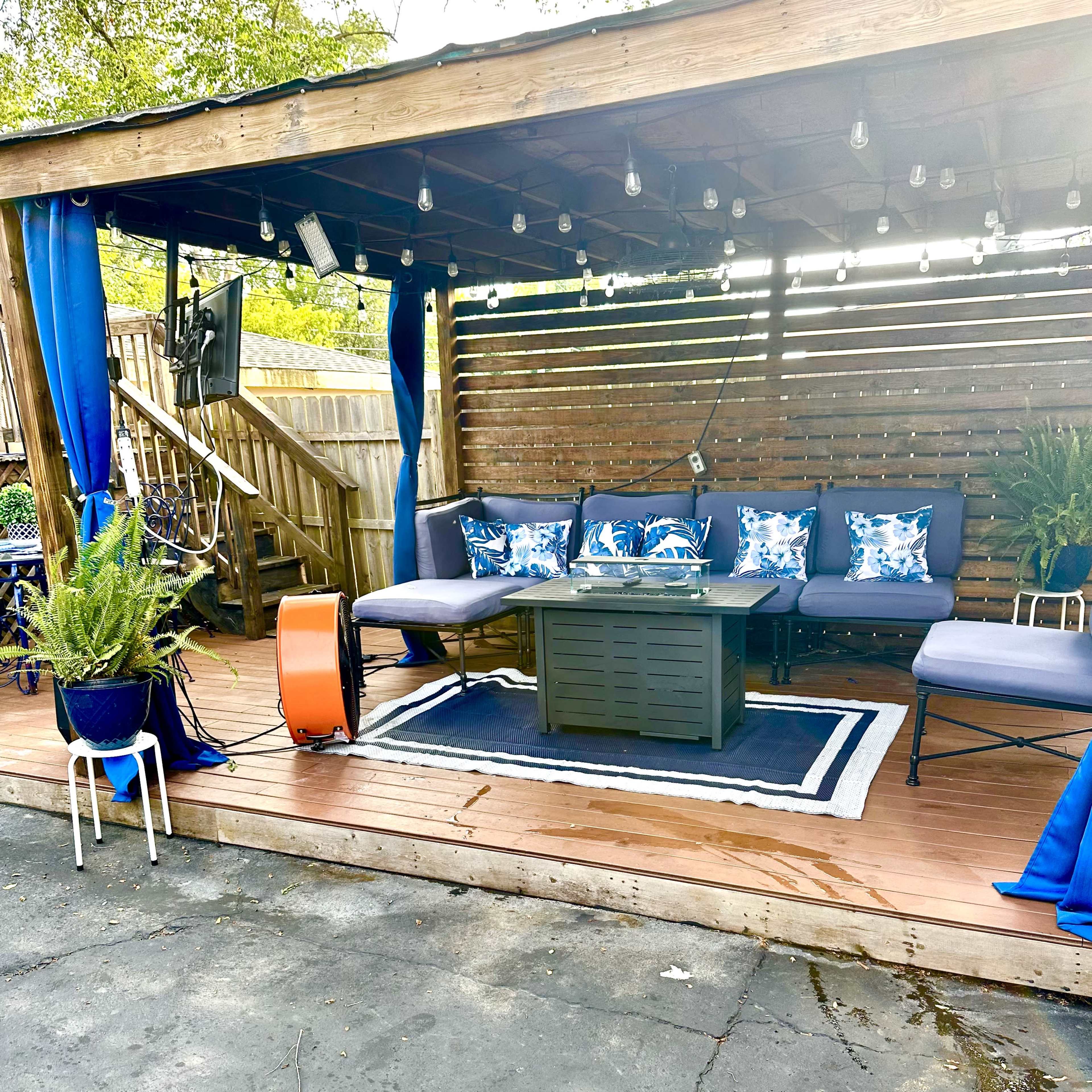 A wooden patio area with a sofa, a coffee table, and decorative pillows, surrounded by string lights and potted plants.