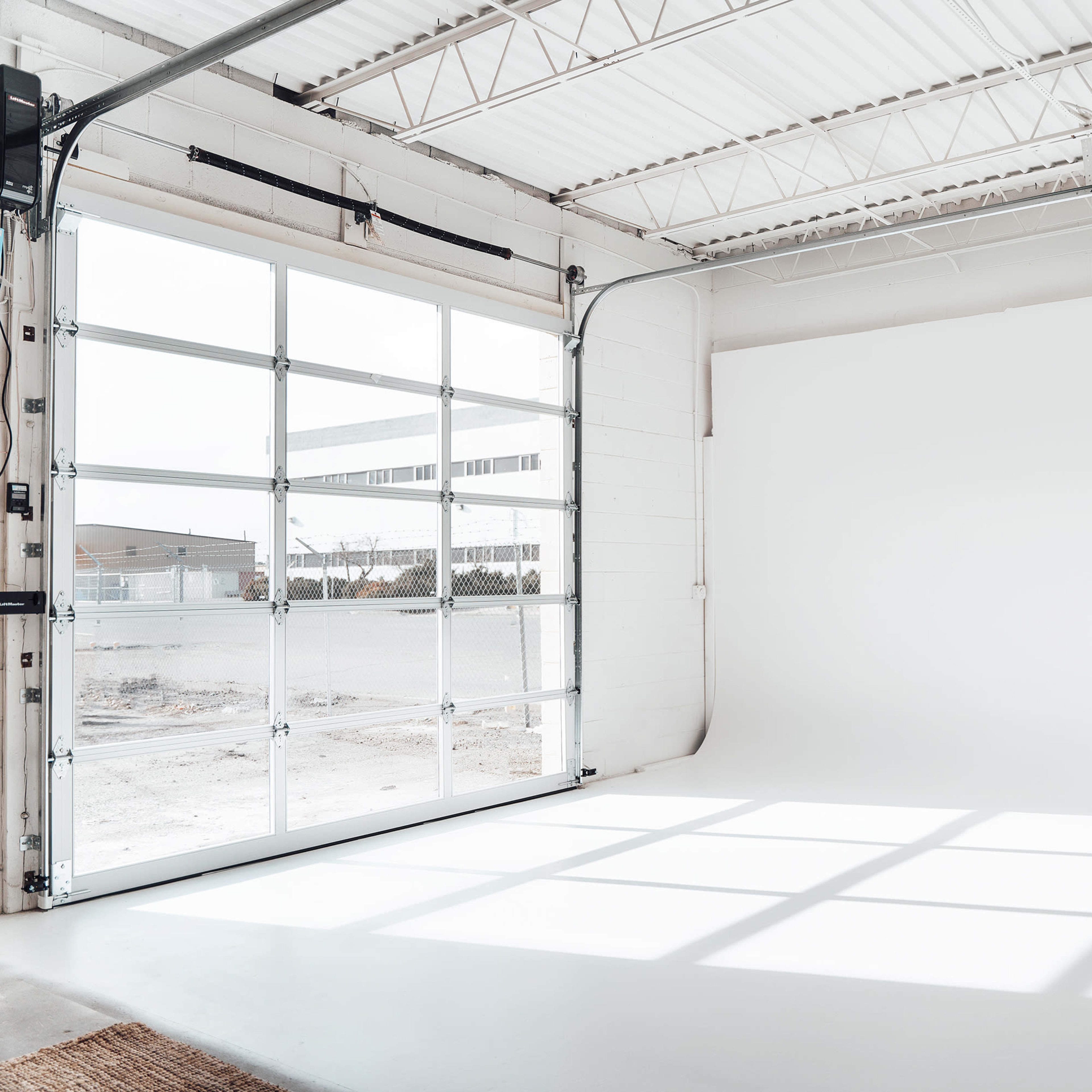 A spacious studio with a large garage door open to a well-lit interior, featuring a plain white backdrop and sunlight casting shadows on the floor.