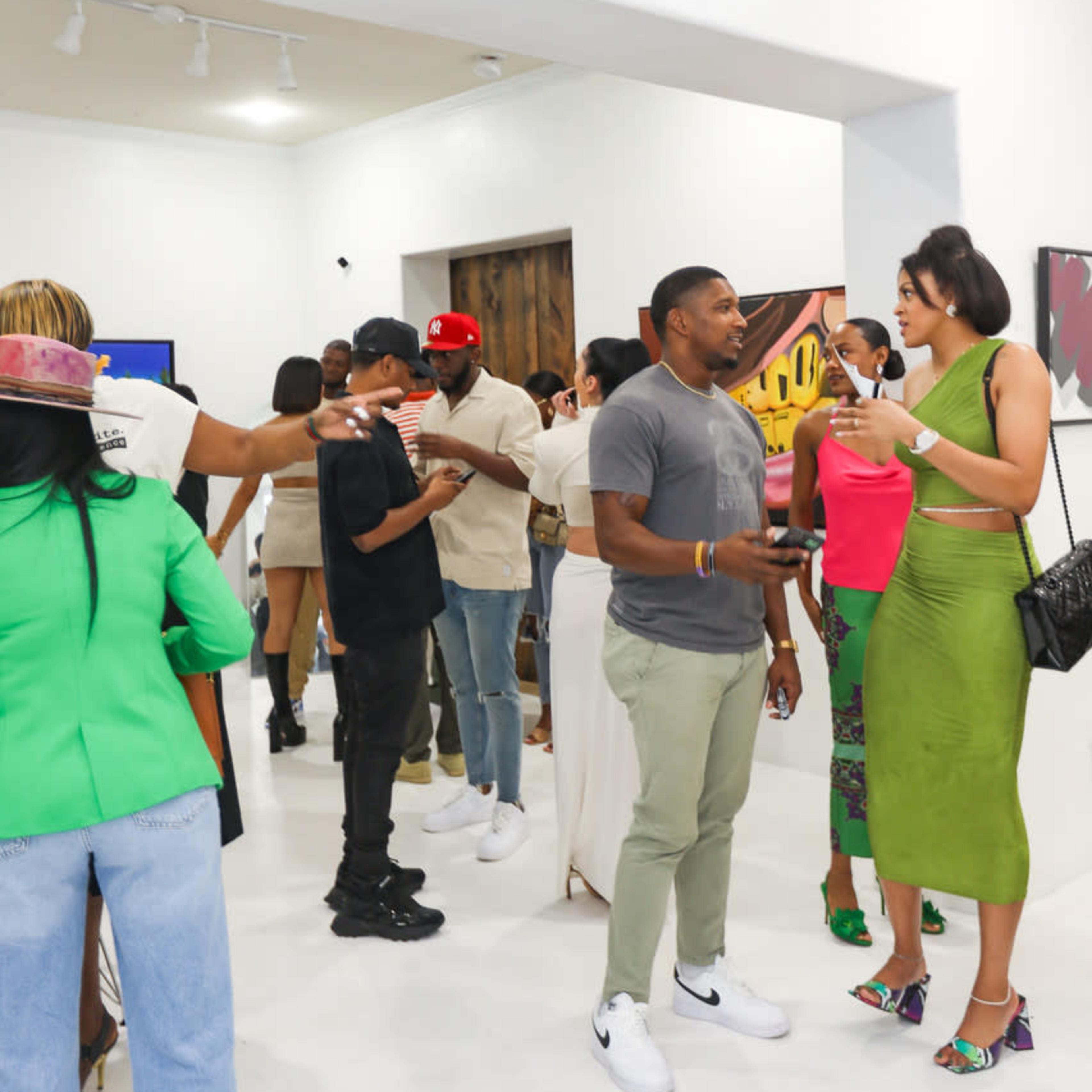 A group of people socialize in an art gallery, engaging in conversation and holding drinks amidst various artworks on display.